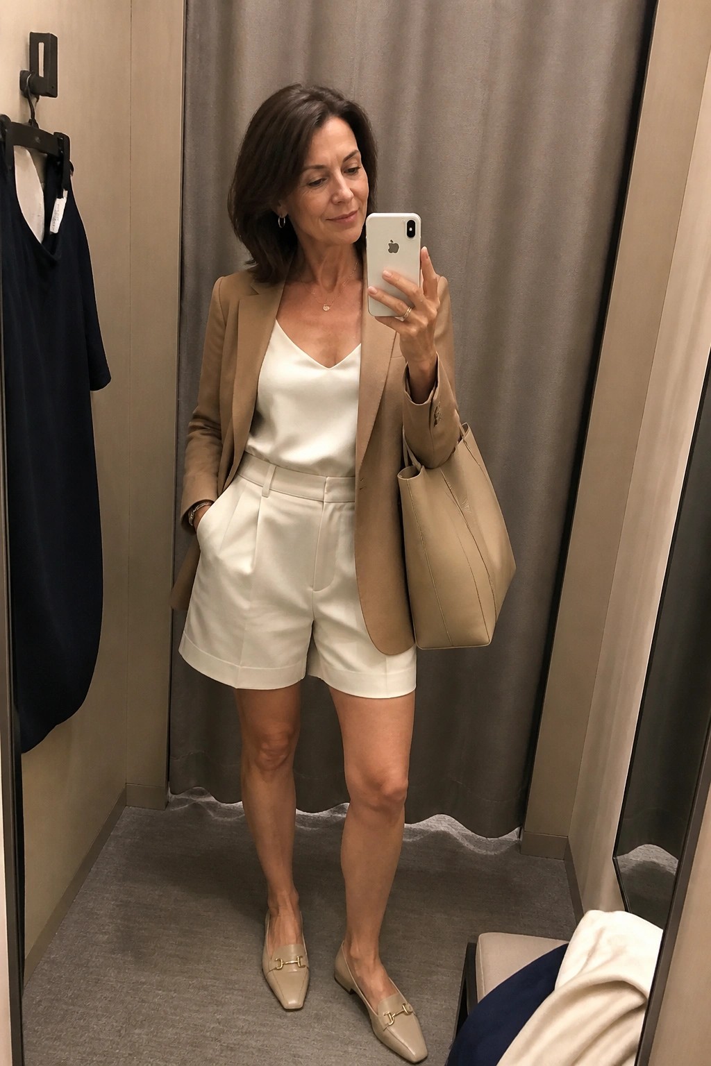 Woman in a camel blazer over a white camisole and white pleated shorts with beige loafers and matching tote bag taking a mirror selfie in a fitting room.