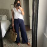 Woman over fifty in white sleeveless linen top tucked into navy wide leg pants with brown woven bag and matching slides holding phone for mirror selfie