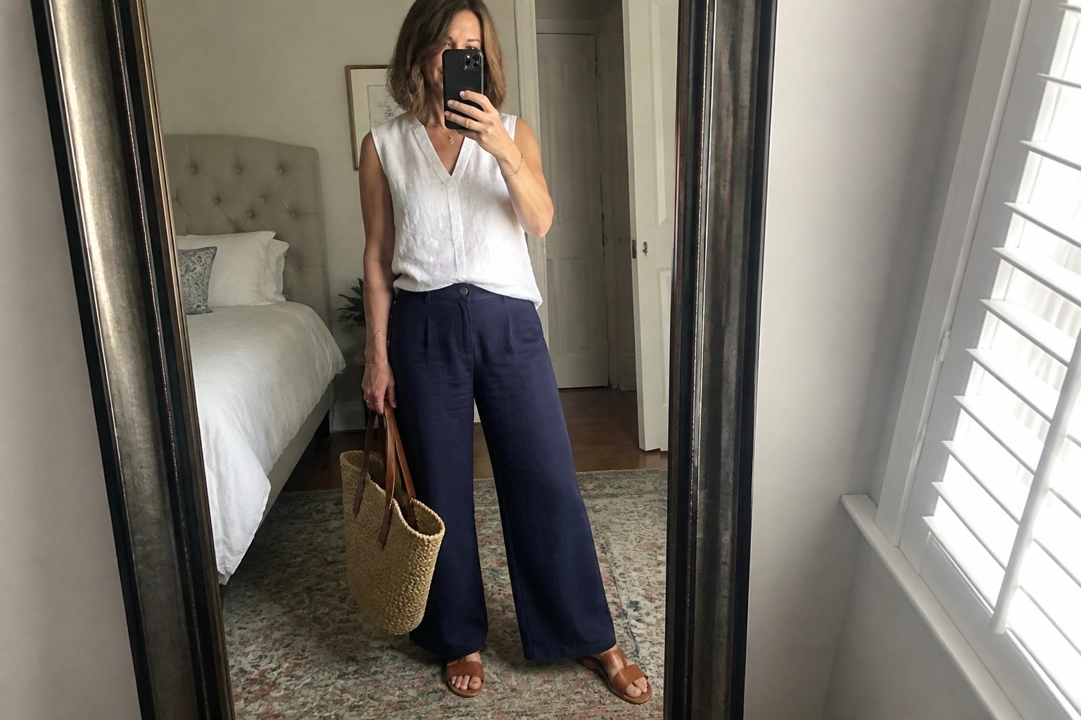 Woman over fifty in white sleeveless linen top tucked into navy wide leg pants with brown woven bag and matching slides holding phone for mirror selfie