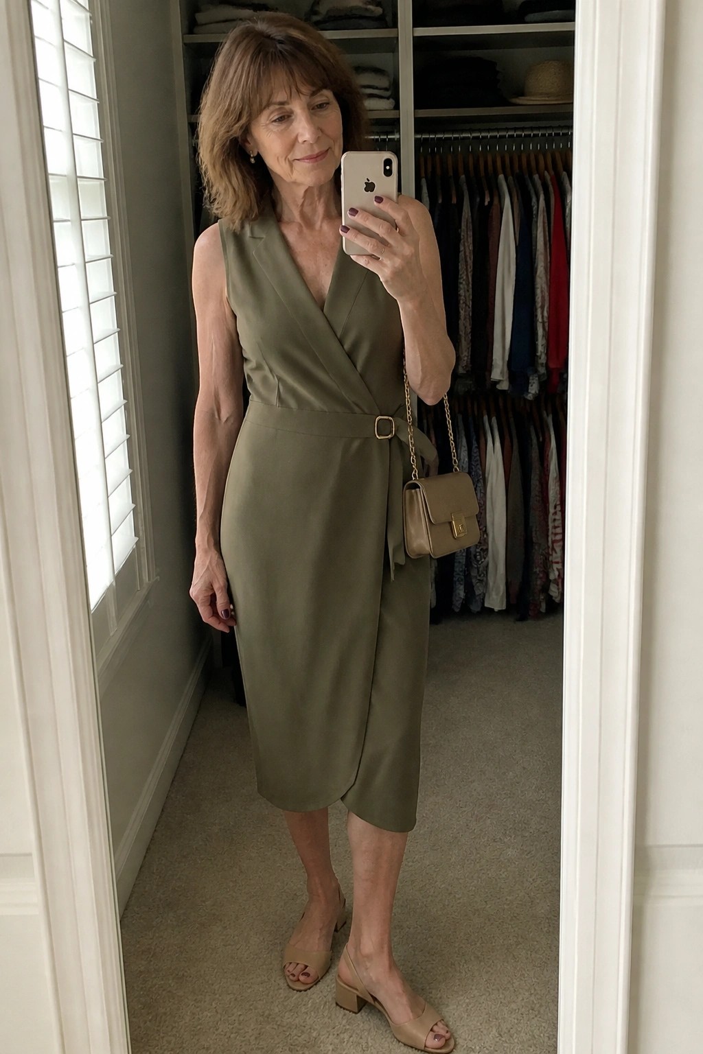 Woman wearing an olive green sleeveless wrap dress with a belted waist, beige chain strap handbag, and matching block heel sandals while taking a mirror selfie.
