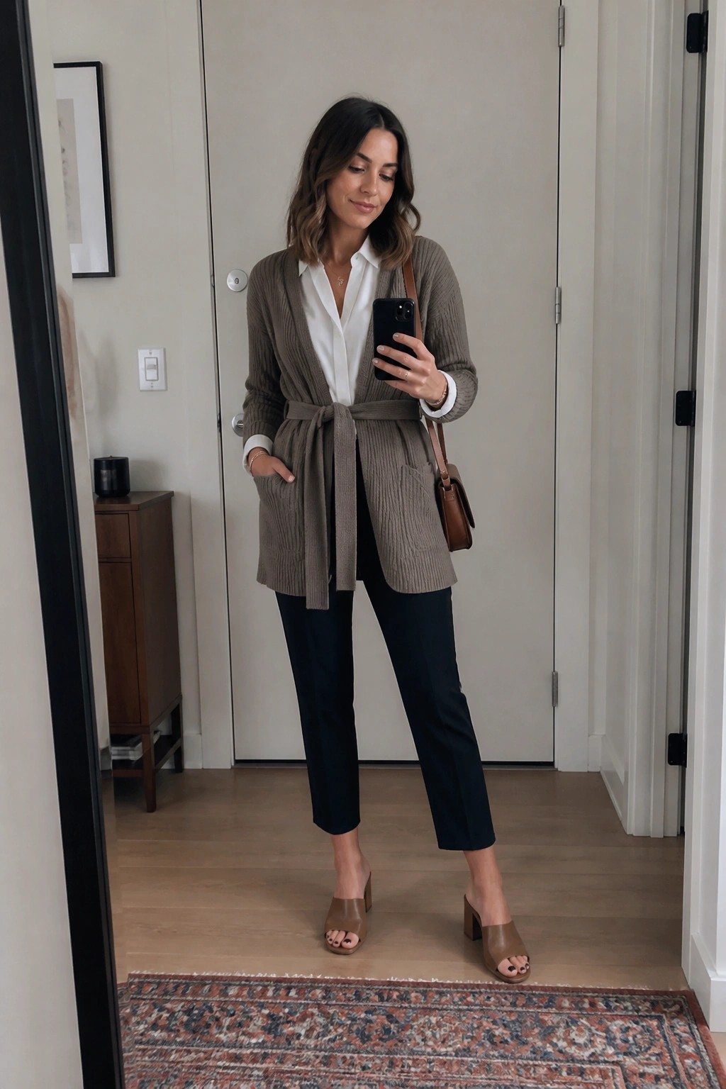 Woman in belted taupe cardigan over white shirt with black pants taking mirror selfie