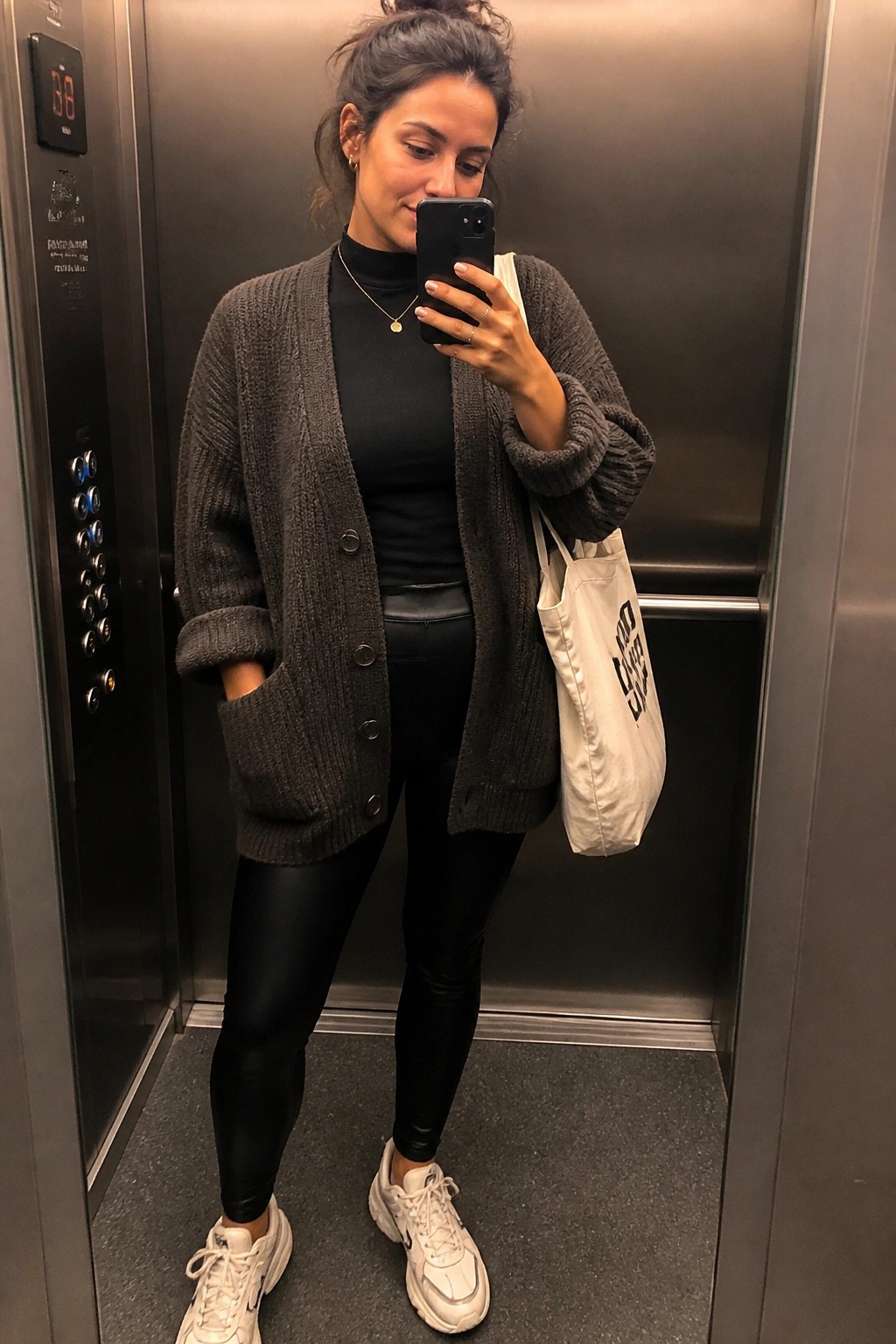 Woman in gray cardigan and black leggings taking selfie in elevator with tote bag