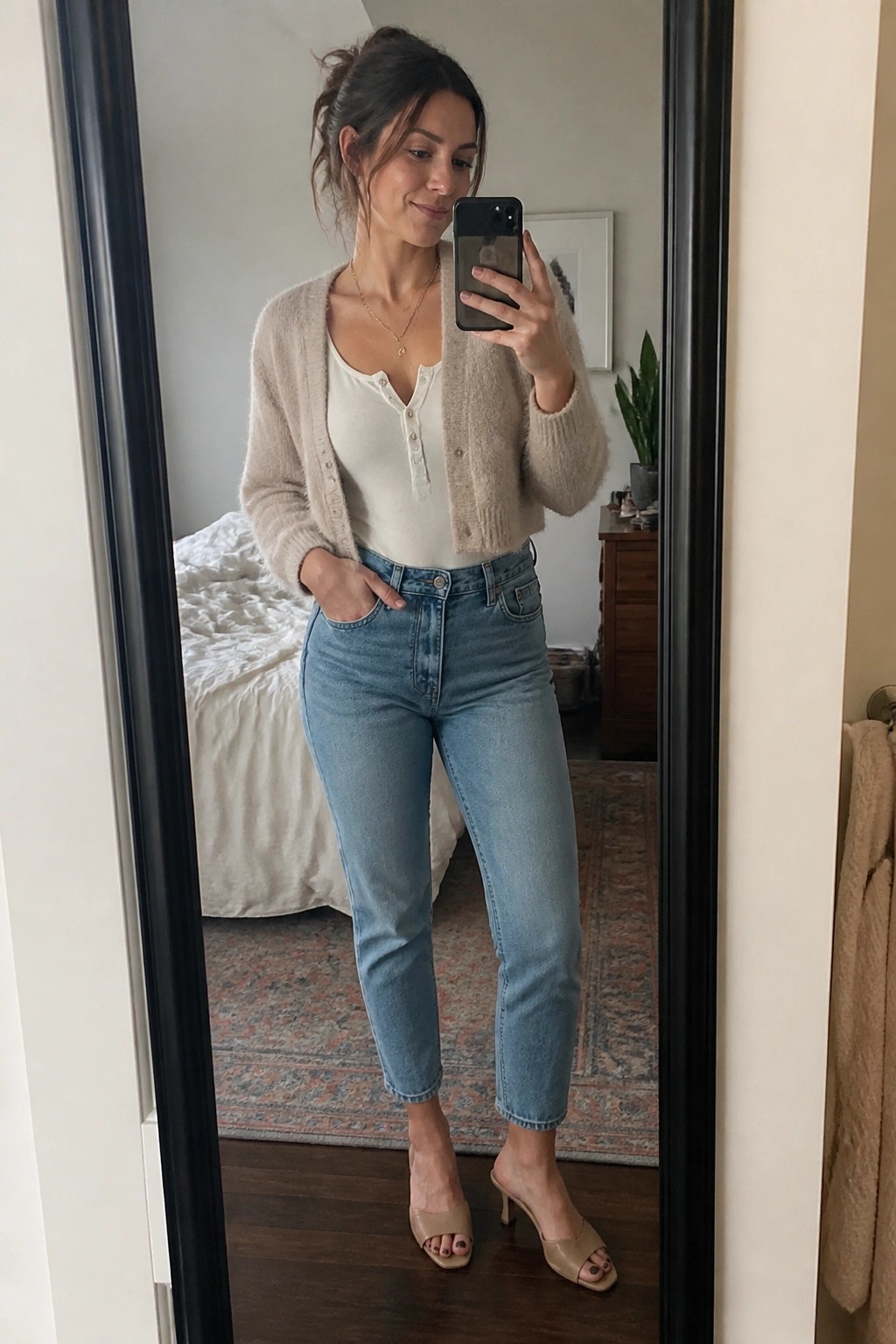 Woman in beige cardigan and jeans taking mirror selfie in bedroom