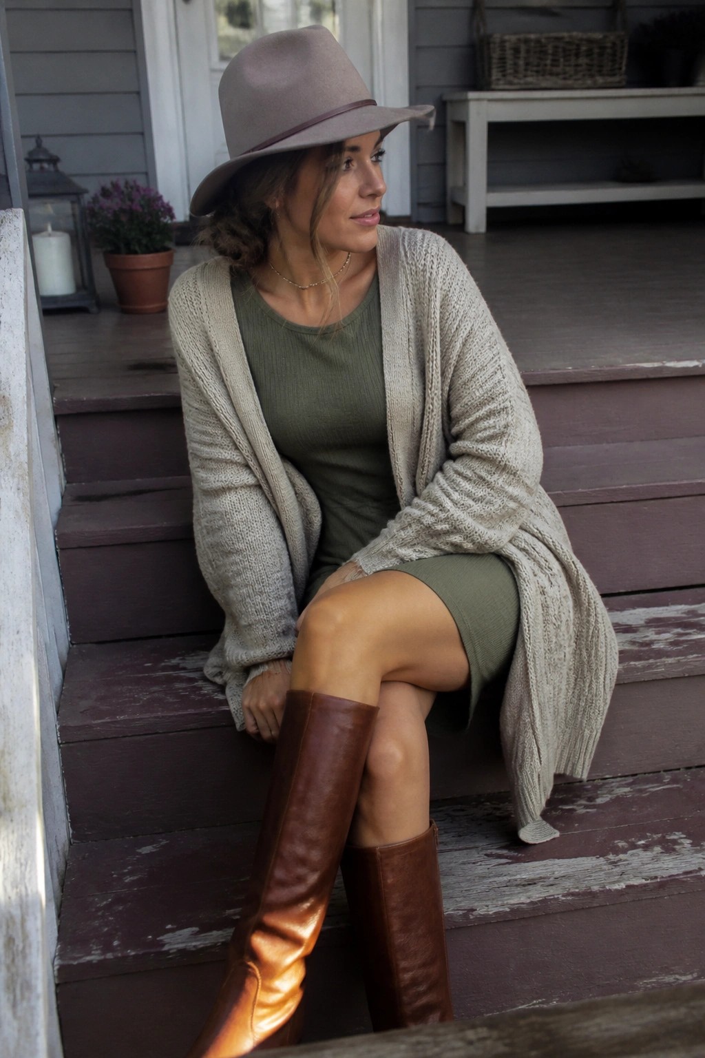 Woman in olive dress, beige cardigan, brown boots, and hat on porch steps