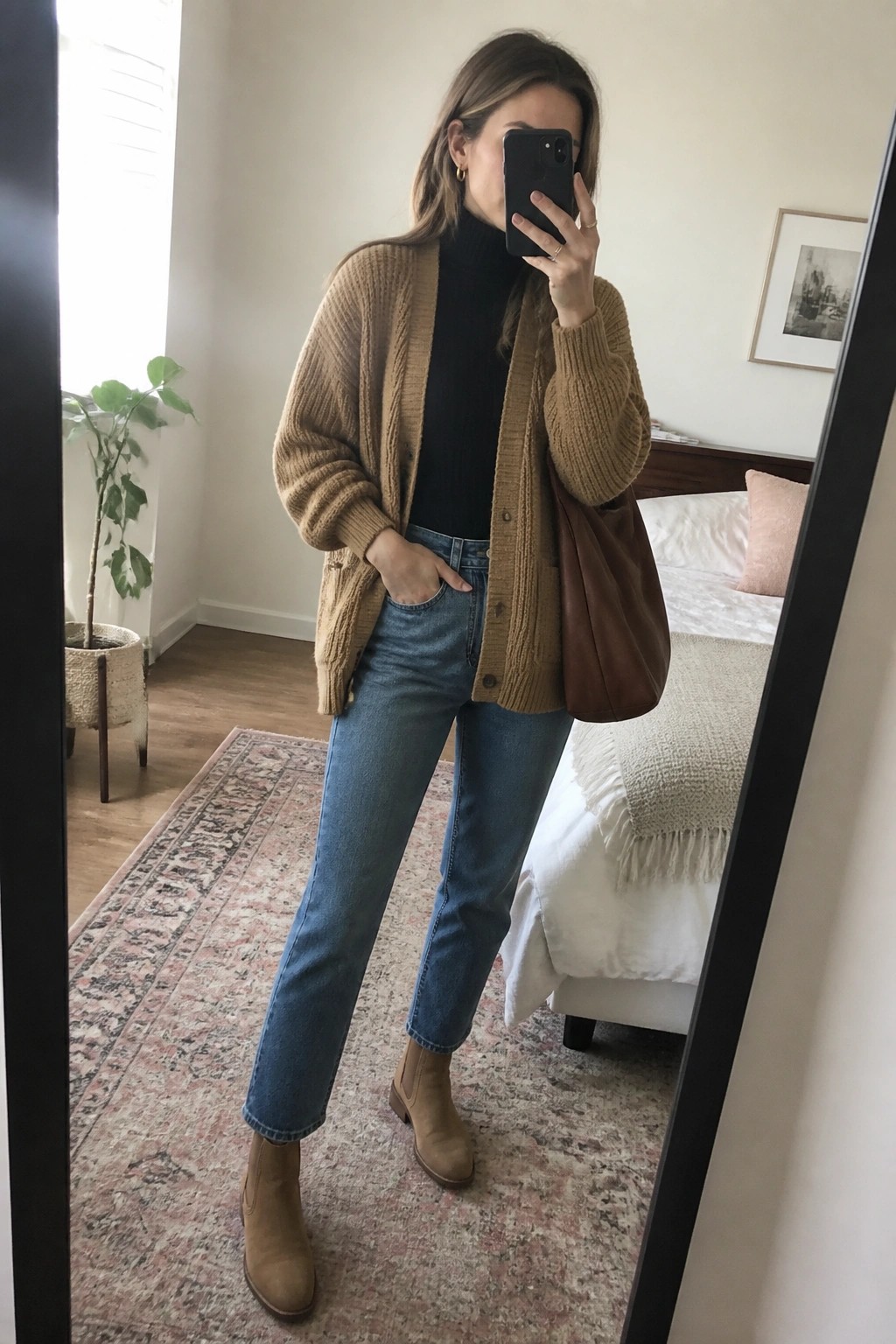 Woman in tan cardigan over black turtleneck taking mirror selfie in bedroom.