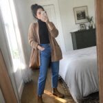 Woman in tan cardigan over black turtleneck taking mirror selfie in bedroom.