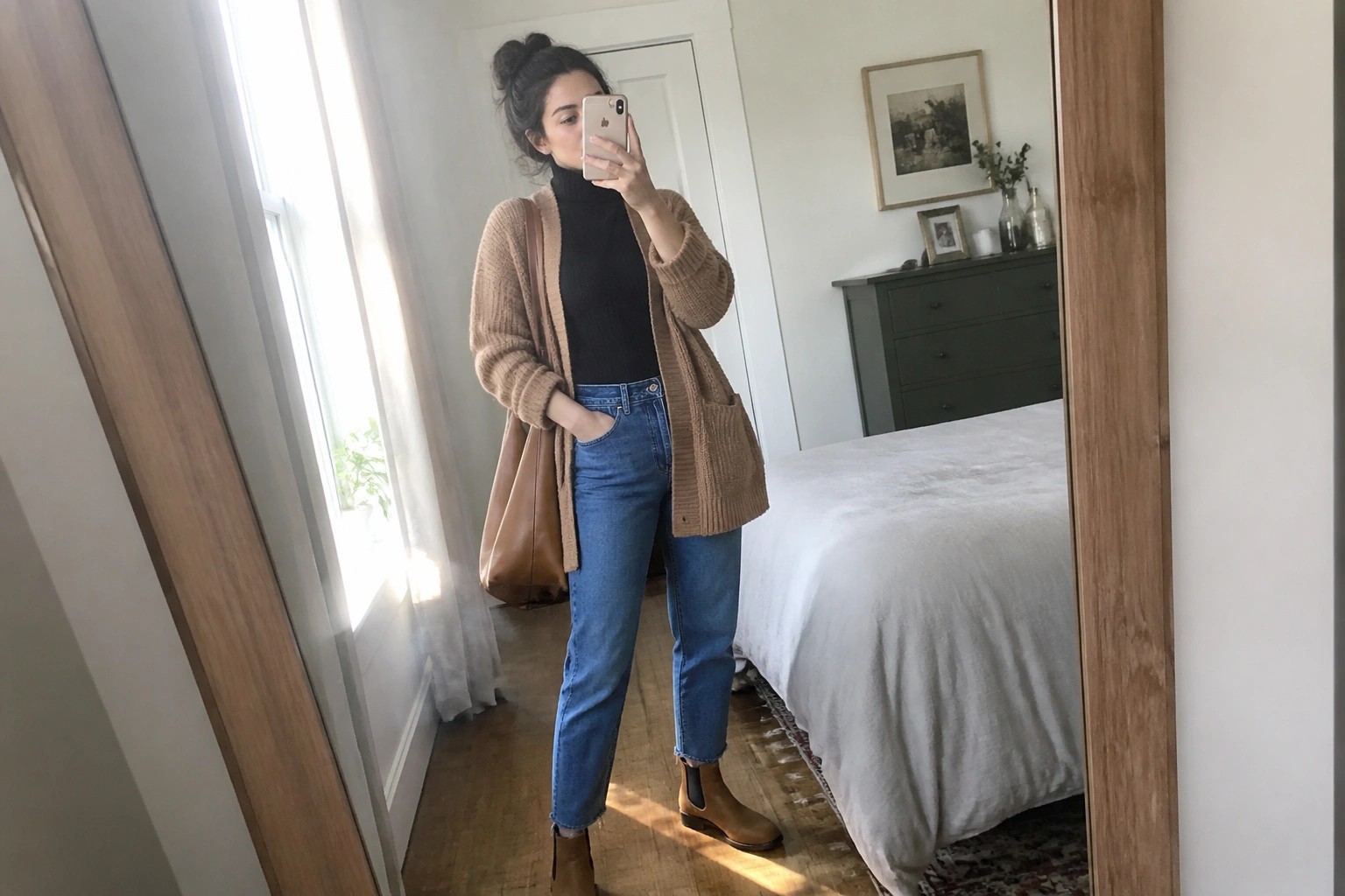 Woman in tan cardigan over black turtleneck taking mirror selfie in bedroom.