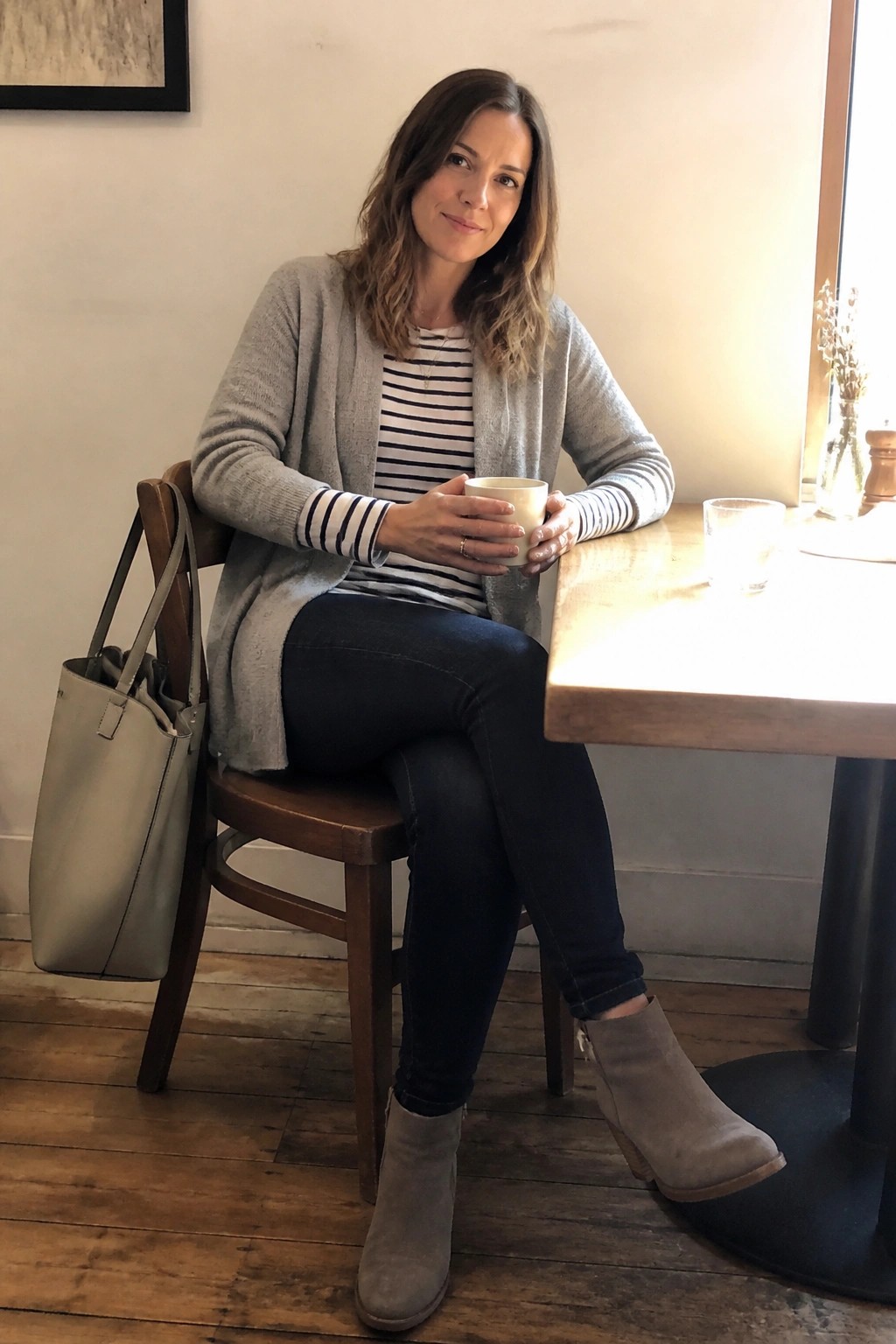 Woman in gray cardigan and striped shirt holding mug at wooden cafe table