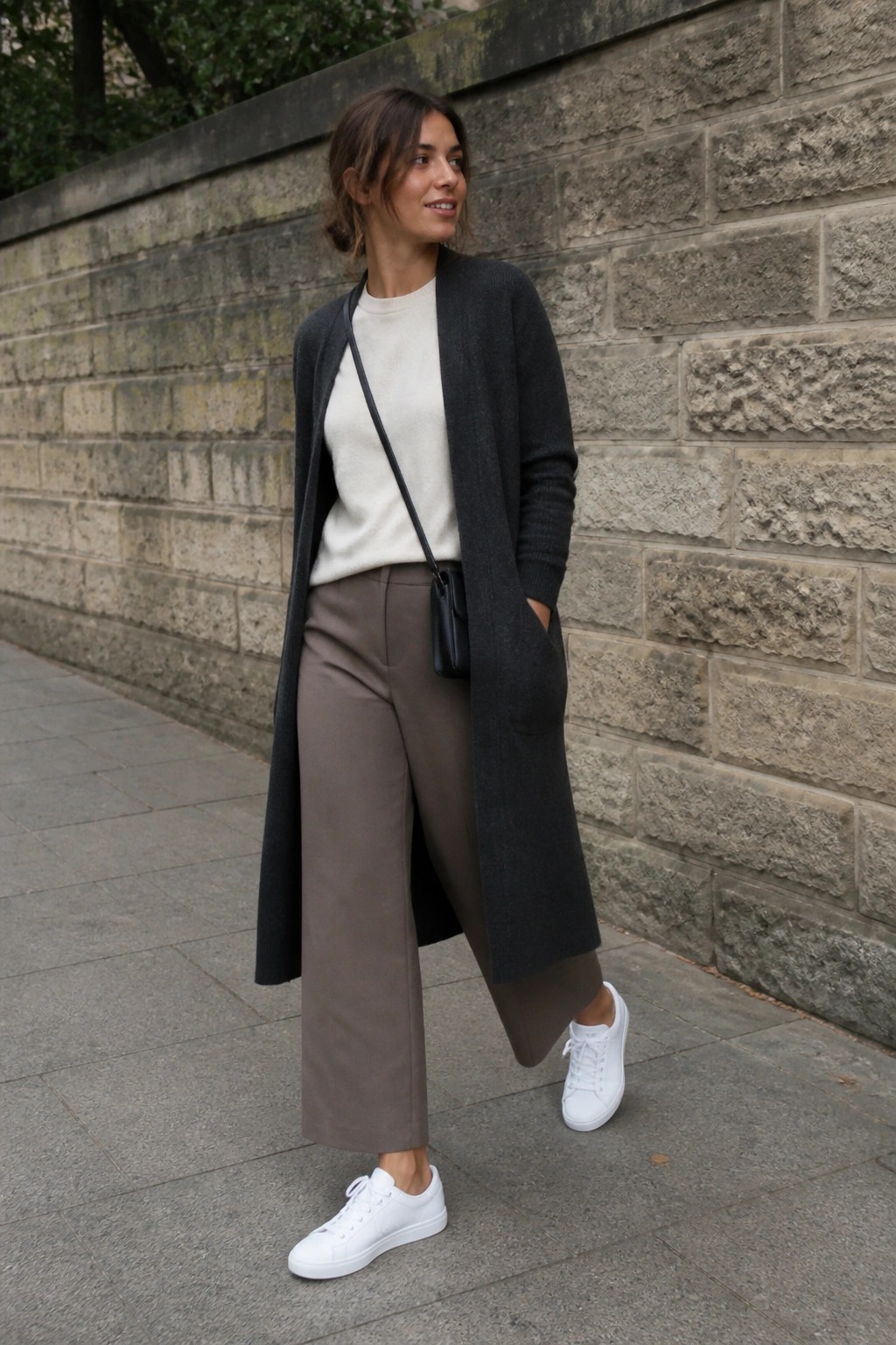 Woman in long dark cardigan, cream sweater, taupe pants, white sneakers by stone wall.