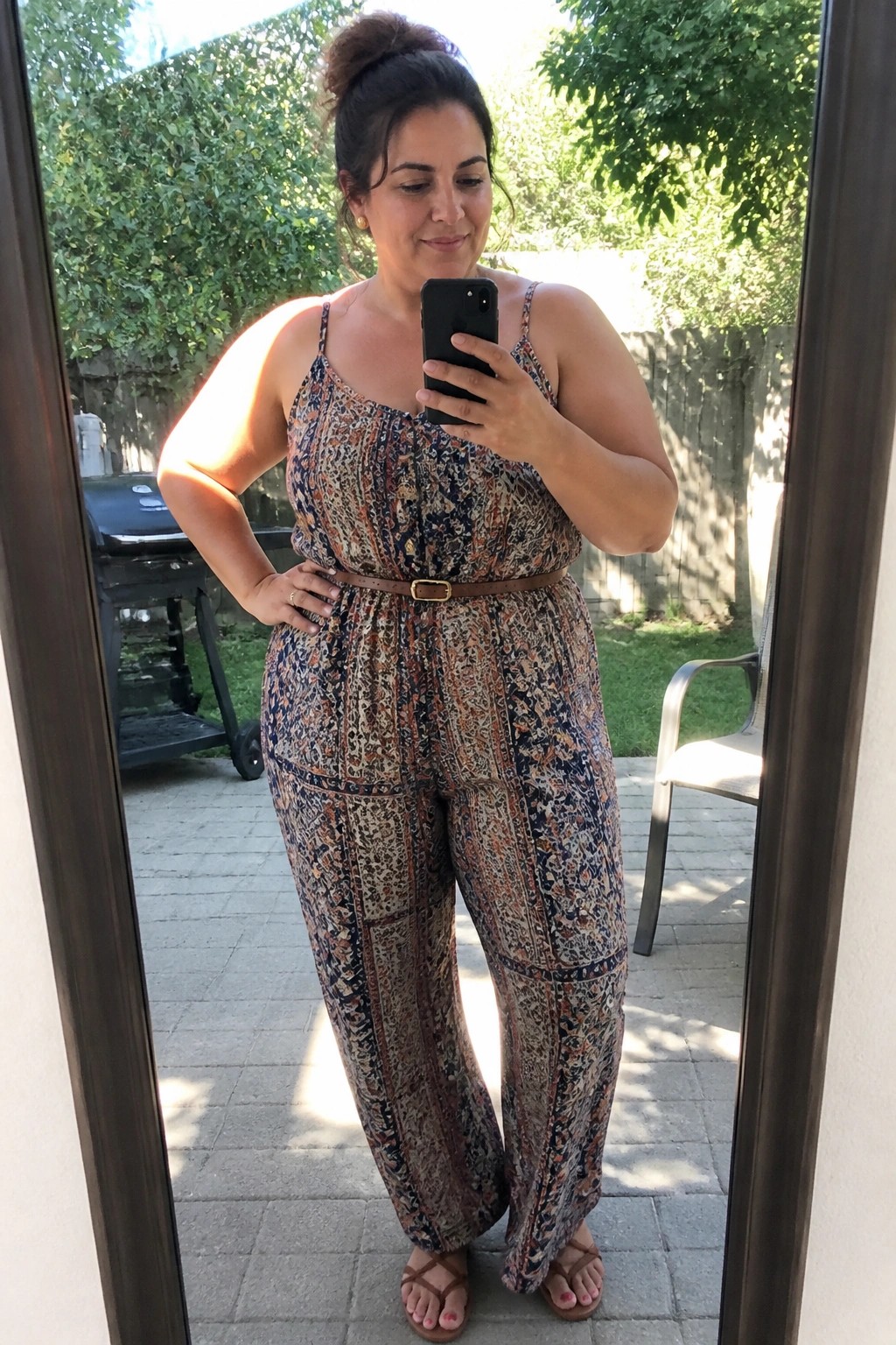 Woman in patterned jumpsuit with belt taking mirror selfie on patio
