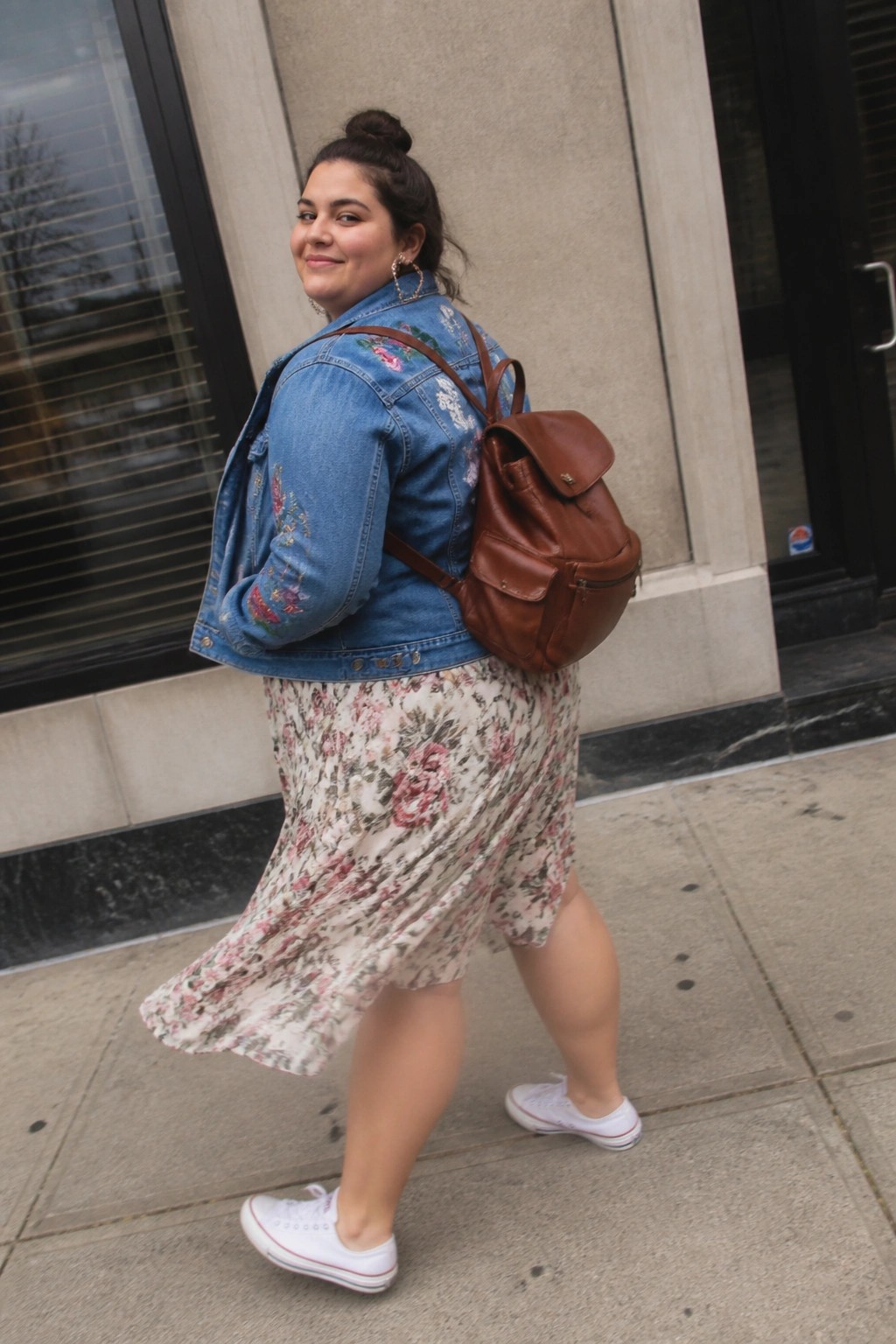 Woman in embroidered denim jacket and floral skirt with brown backpack on sidewalk