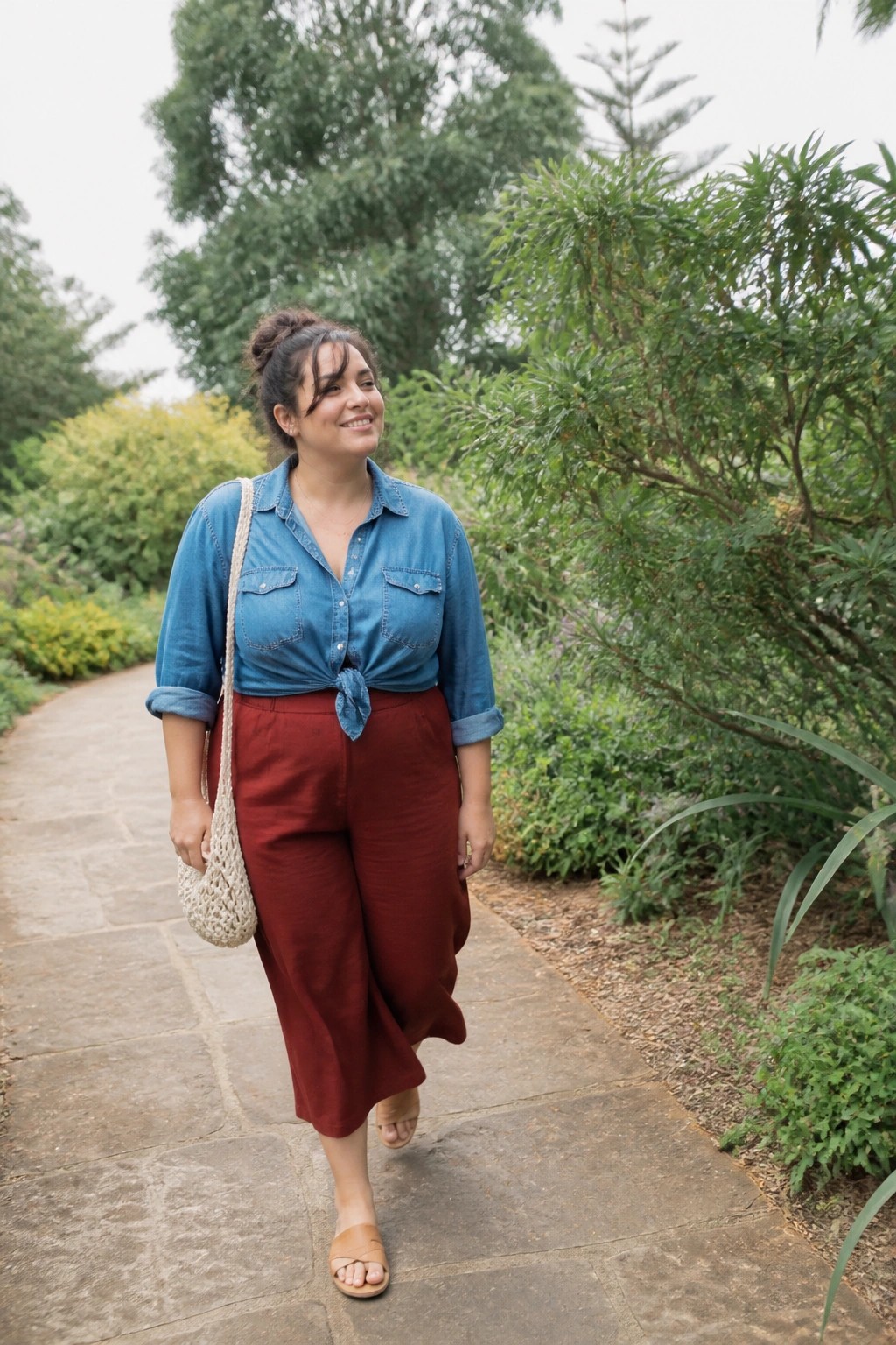 Woman in blue denim shirt and red pants walking on garden path with macrame bag