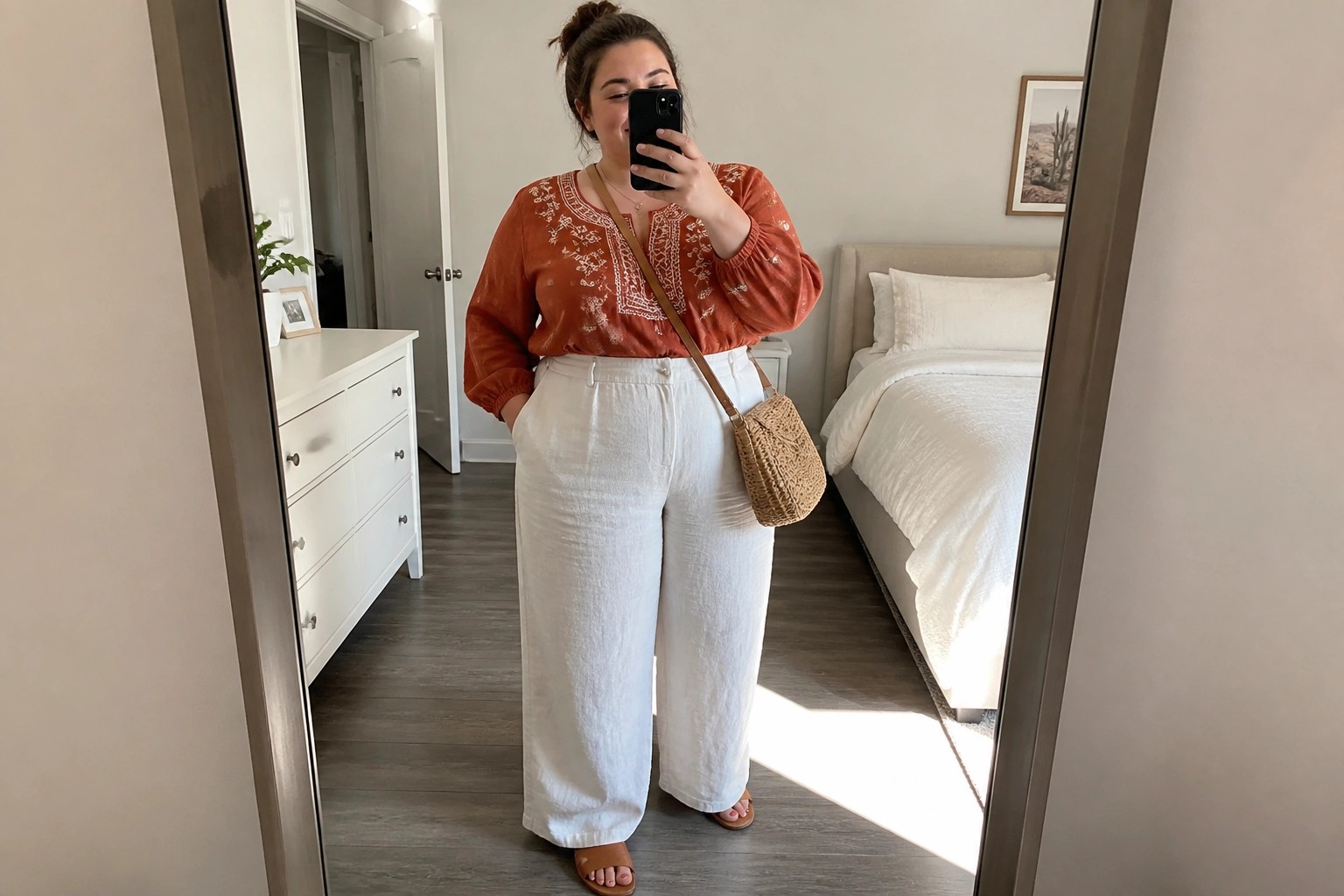 Plus size woman wearing a rust embroidered blouse with wide beige linen pants, round woven crossbody bag, and brown slides while taking a mirror selfie.