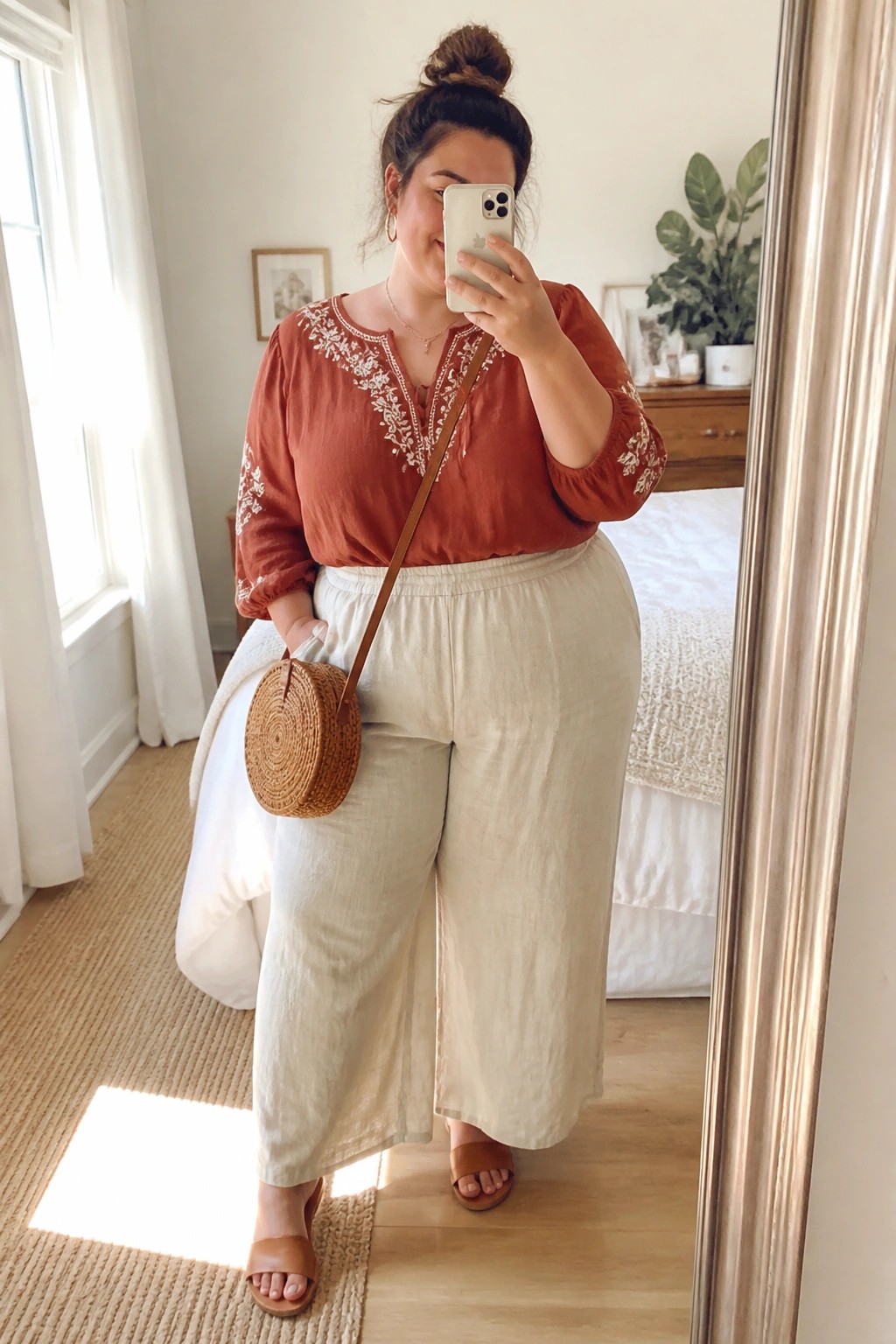 Plus size woman wearing a rust embroidered blouse with wide beige linen pants, round woven crossbody bag, and brown slides while taking a mirror selfie.