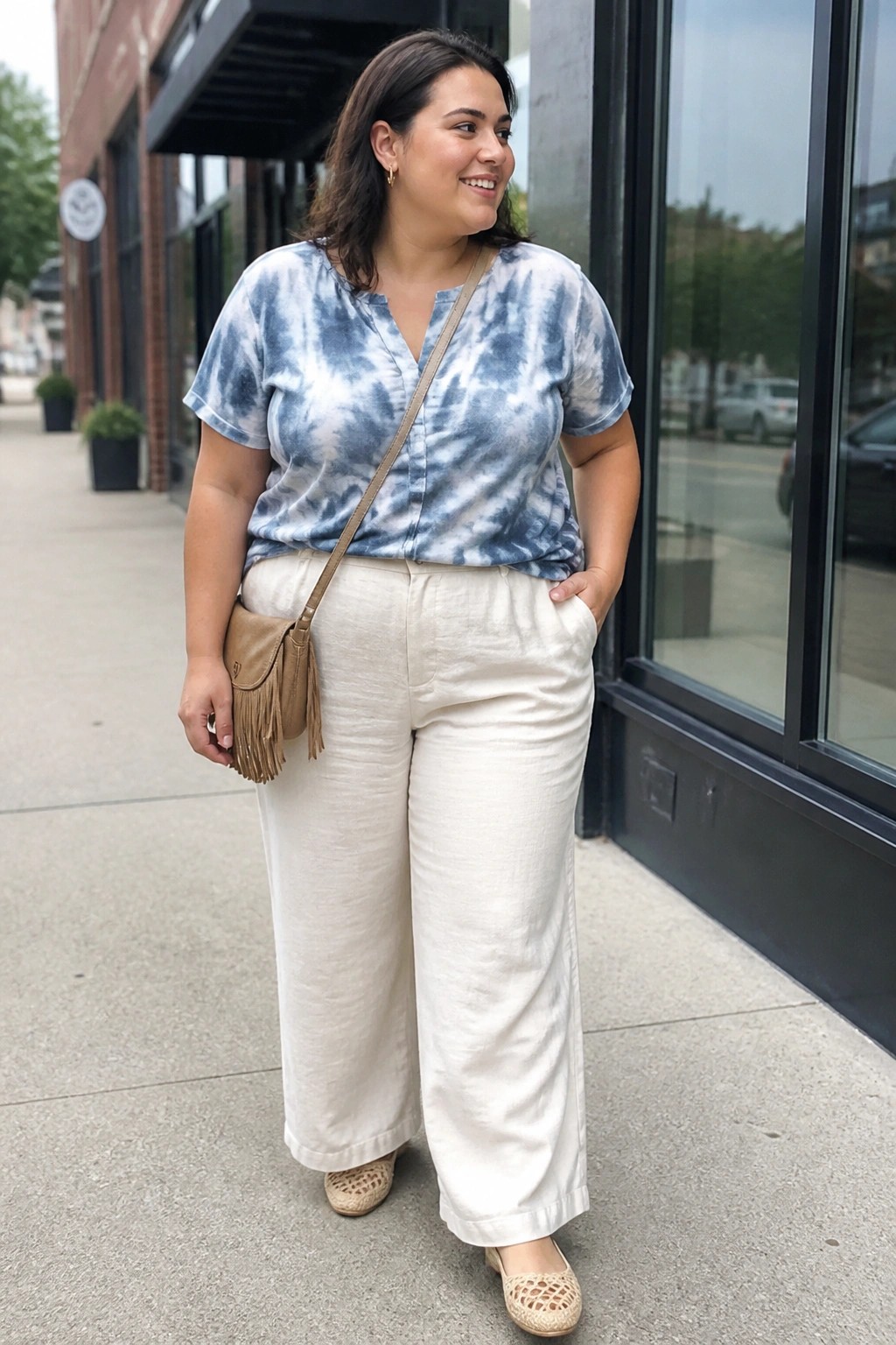 Woman in blue tie-dye top and white wide-leg pants with fringed bag on sidewalk