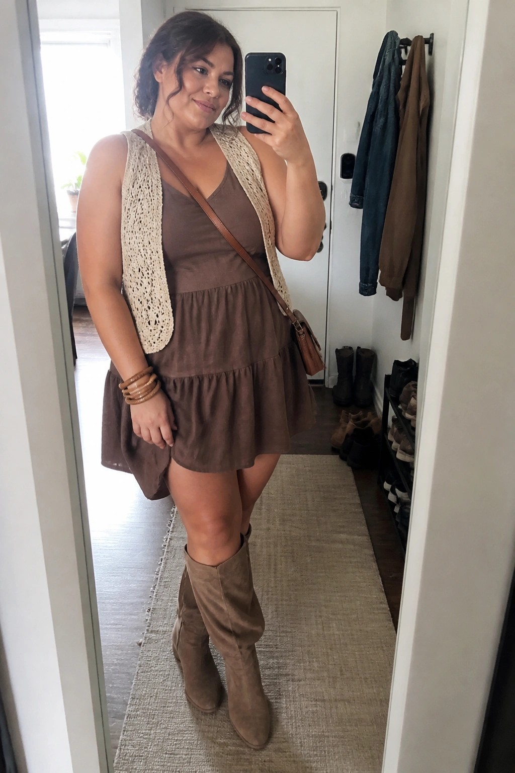 Woman wearing a brown tiered mini dress under a cream open crochet vest, paired with matching suede knee high boots, a brown crossbody bag, and stacked wooden bangles while taking a mirror selfie.