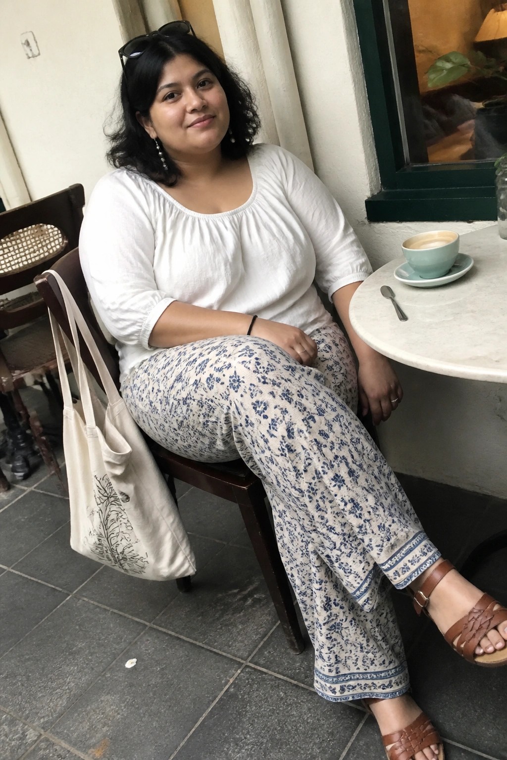 A plus size woman in a white scoop neck top and blue floral wide leg pants sitting at a cafe table with a tote bag.