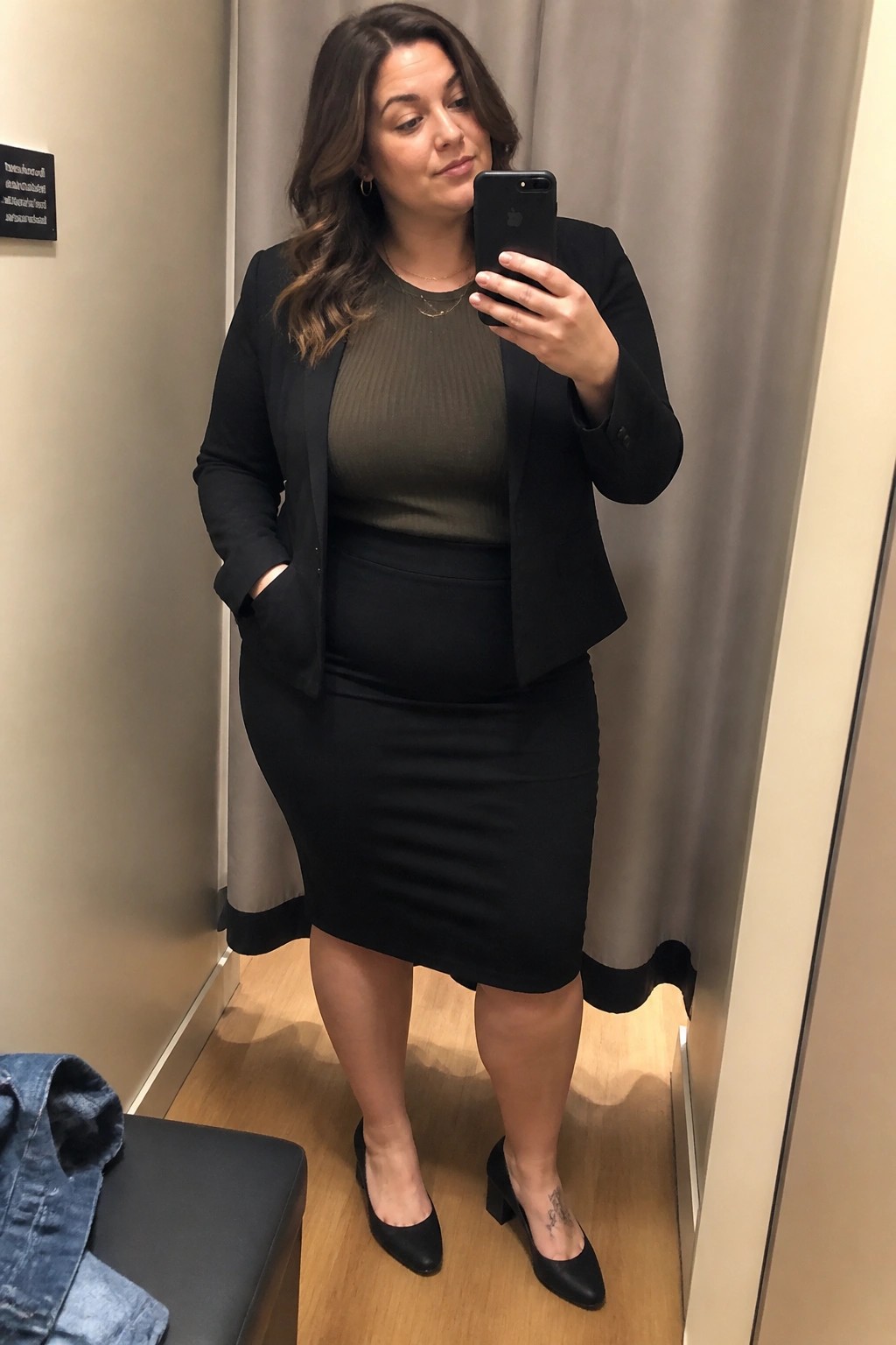 Woman in black blazer and skirt taking mirror selfie in fitting room