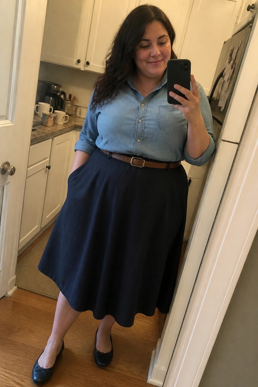 Woman in light blue denim shirt and navy skirt taking mirror selfie in kitchen
