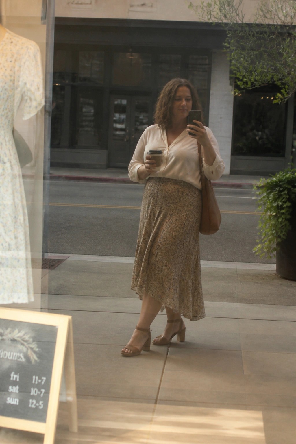 Woman in cream blouse and floral skirt holds coffee while using phone on sidewalk