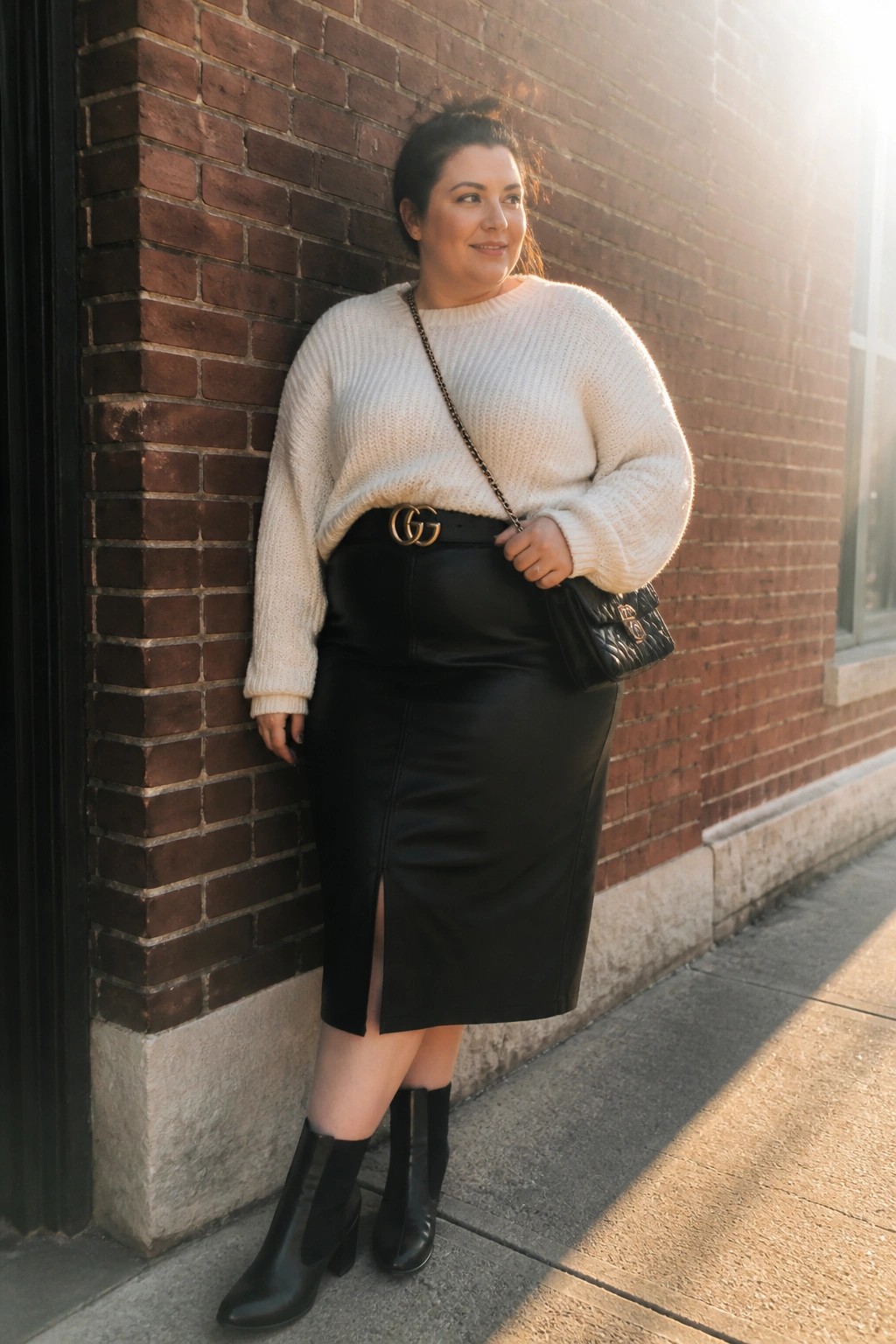Curvy woman in cream knit sweater and black leather skirt against brick wall