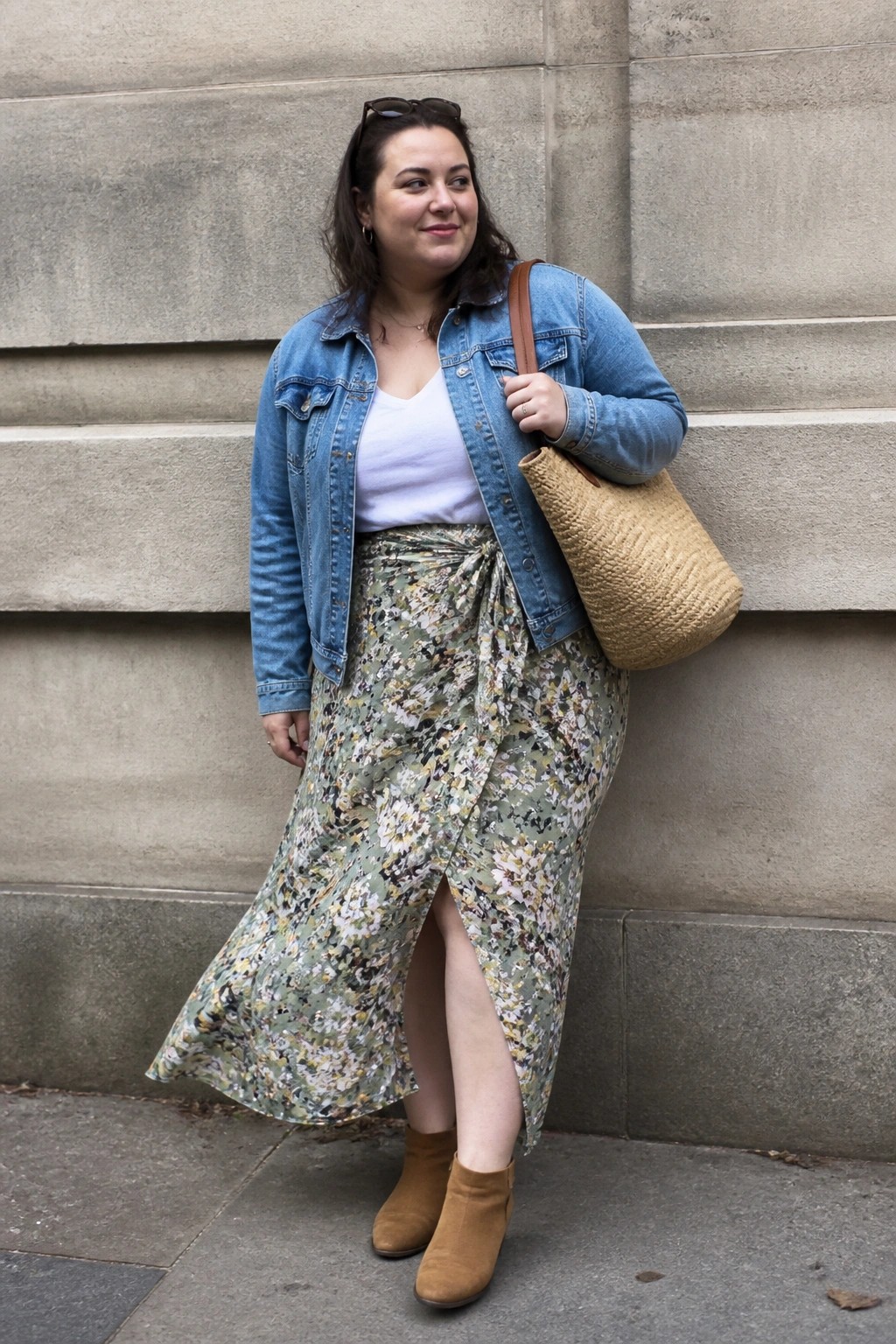 Plus size woman wearing a blue denim jacket over a white tank top paired with a long green floral wrap skirt, brown suede ankle boots, and a woven straw bag.