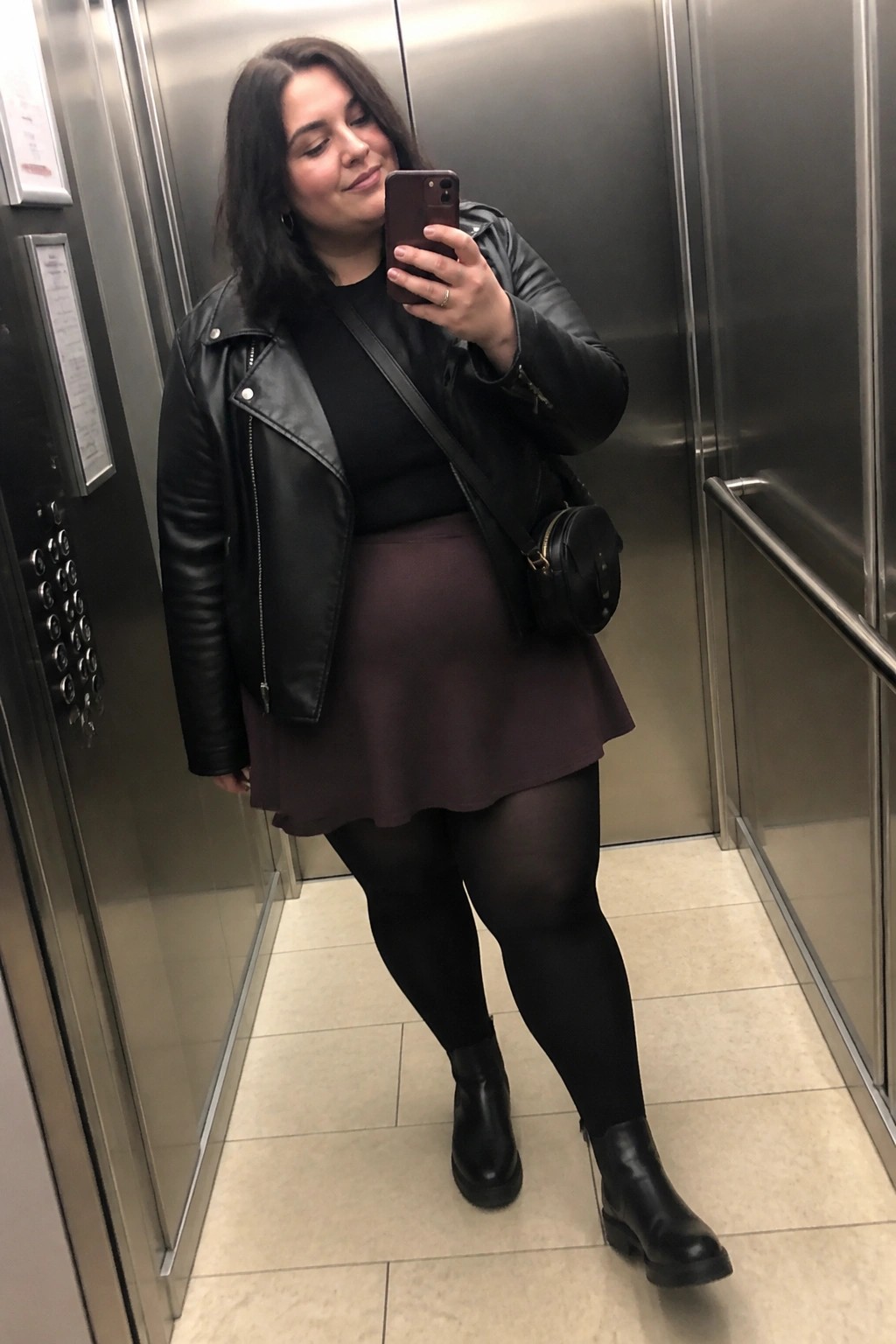 Woman in black leather jacket and maroon skirt taking elevator selfie with phone