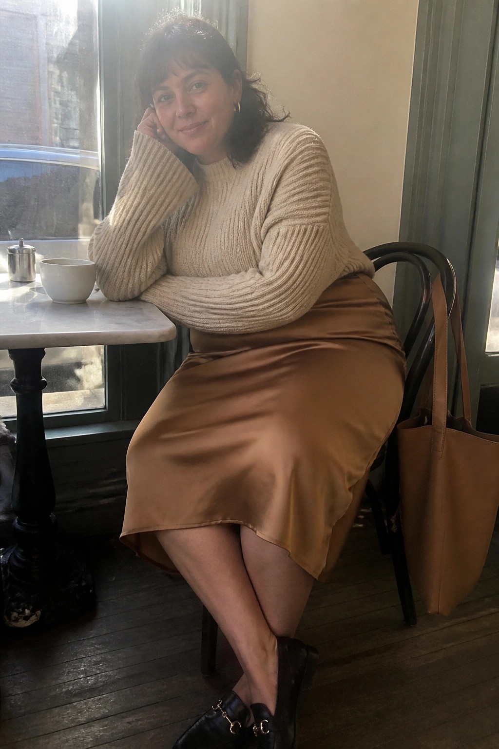 Woman in beige sweater and brown satin skirt seated at cafe table with tote bag