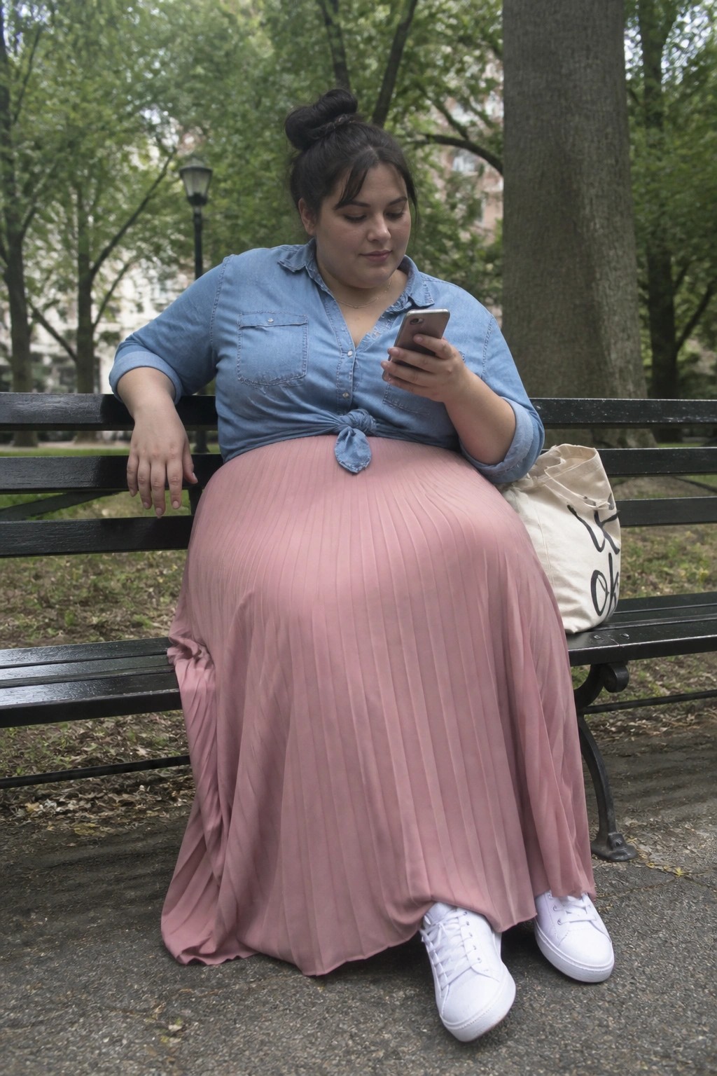 Woman in blue denim shirt and pink pleated skirt uses phone on park bench
