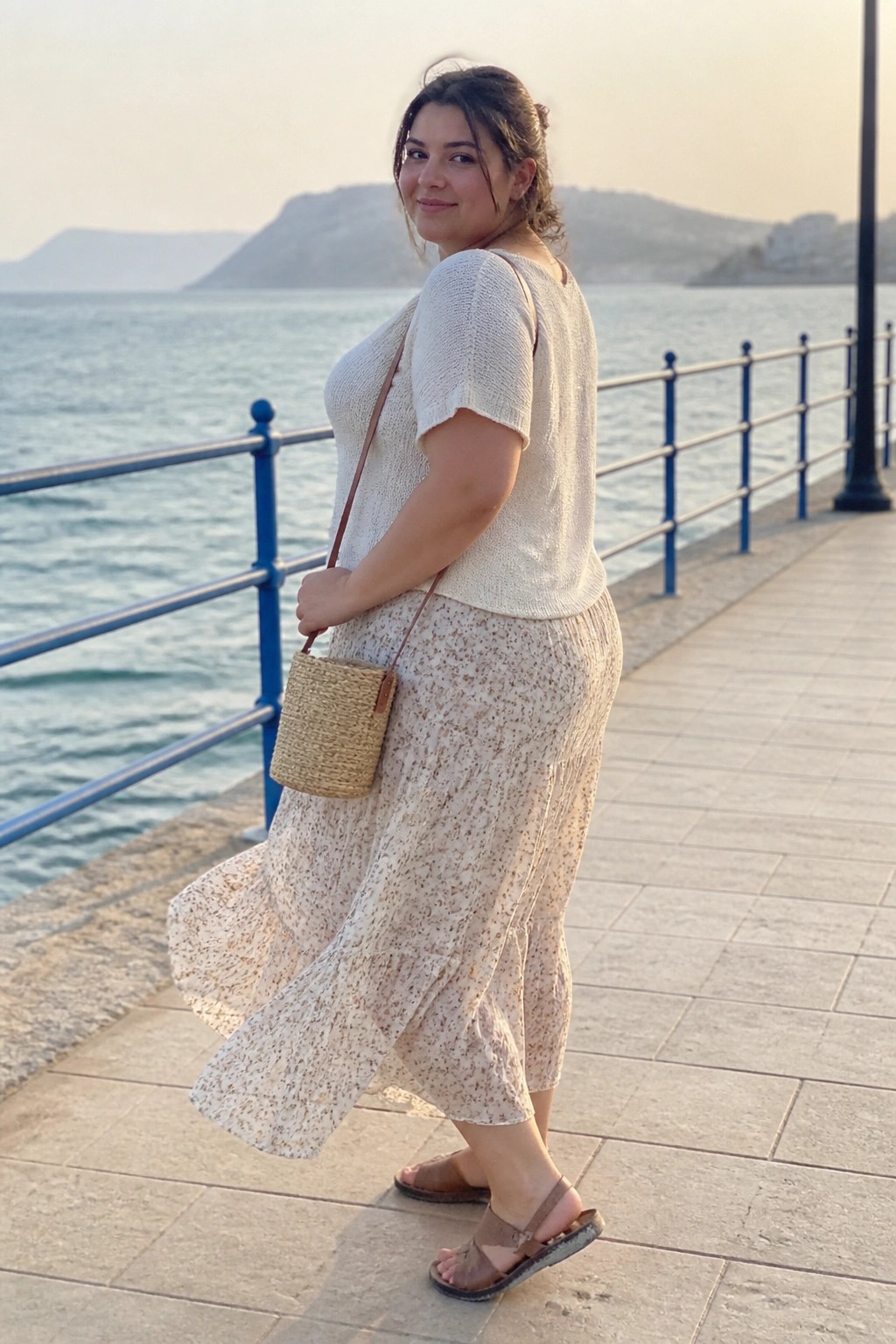 Woman in white knit top and floral skirt with straw bag by the sea.