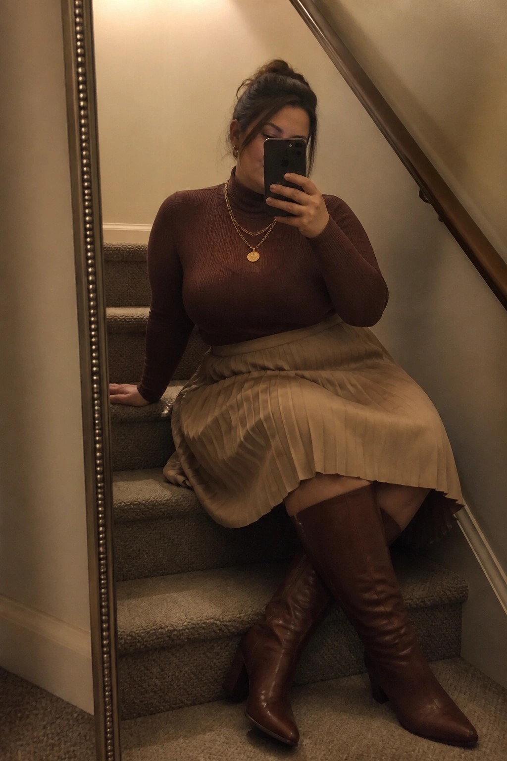 Woman in brown turtleneck and beige pleated skirt taking mirror selfie on stairs