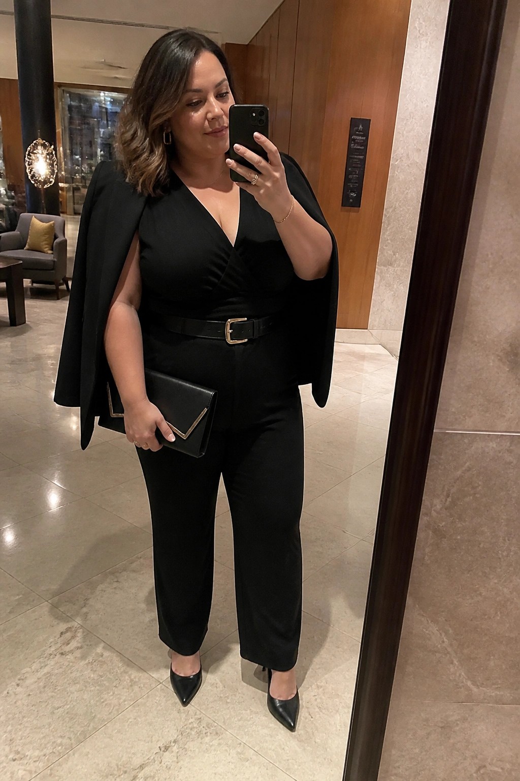 Woman in black jumpsuit with cape jacket taking mirror selfie in elegant lobby