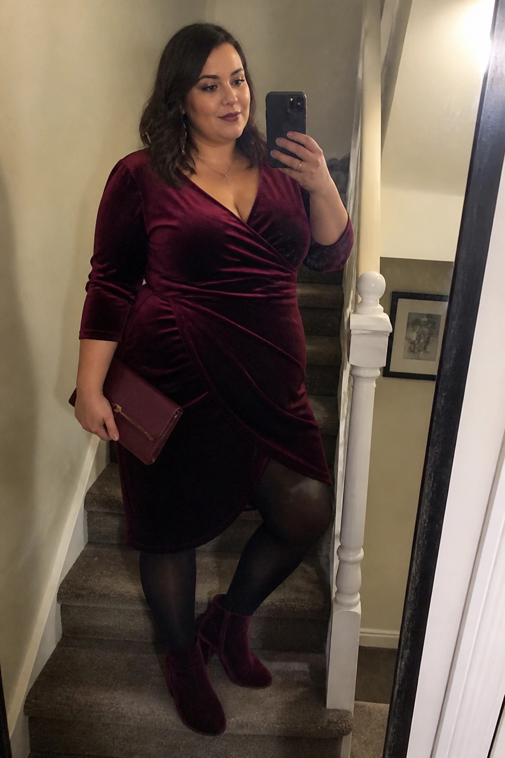 Plus size woman wearing a burgundy velvet wrap dress with three quarter sleeves black tights and matching velvet ankle boots while holding a small burgundy clutch on a staircase.