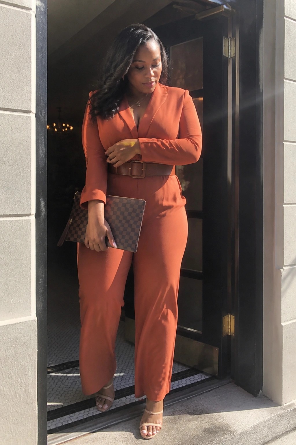 Woman in orange pantsuit holding checkered clutch in sunny doorway