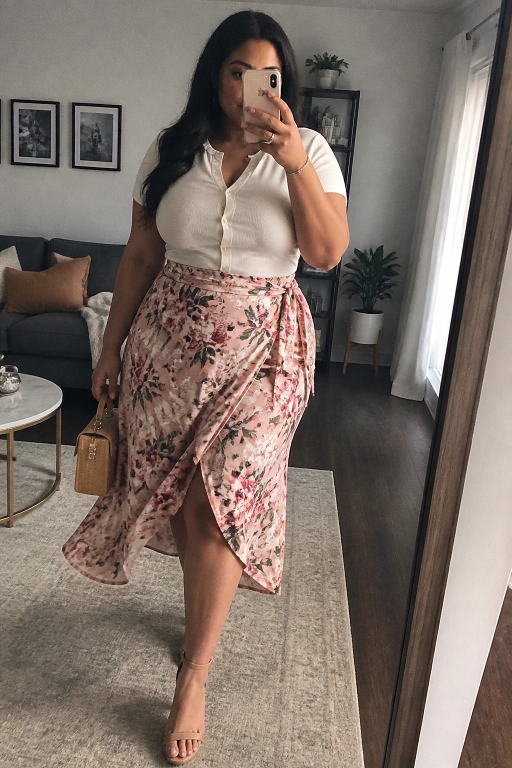 Plus size woman in a white button front top and pink floral wrap skirt holding a beige handbag while taking a mirror selfie in a living room.