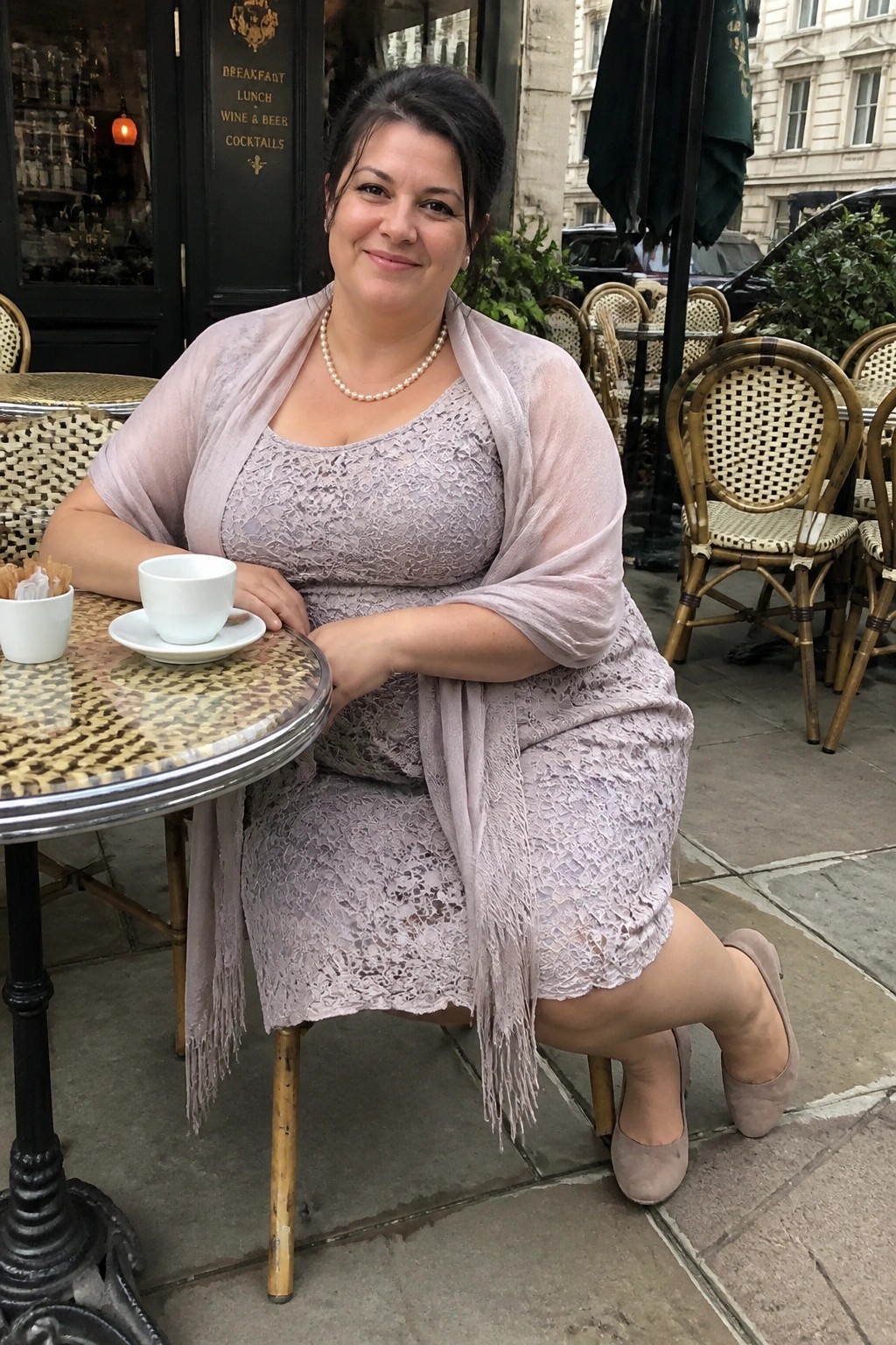 Woman in lavender lace dress seated at outdoor cafe table with coffee cup