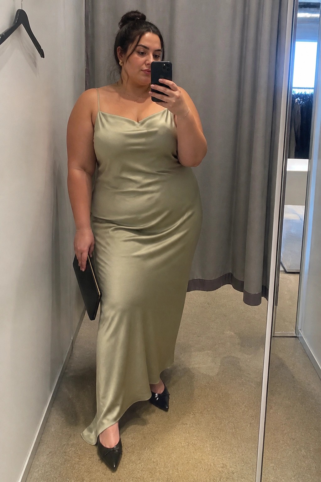 Woman in sage green satin dress taking mirror selfie in fitting room.