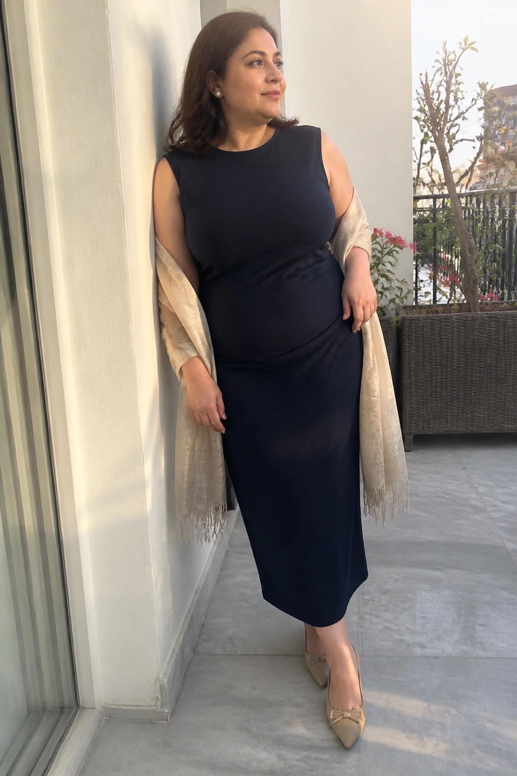 Plus size woman wearing a sleeveless navy midi sheath dress with draped waist detail, light beige patterned fringed shawl over one shoulder, and beige pointed toe heels with small bow accents.