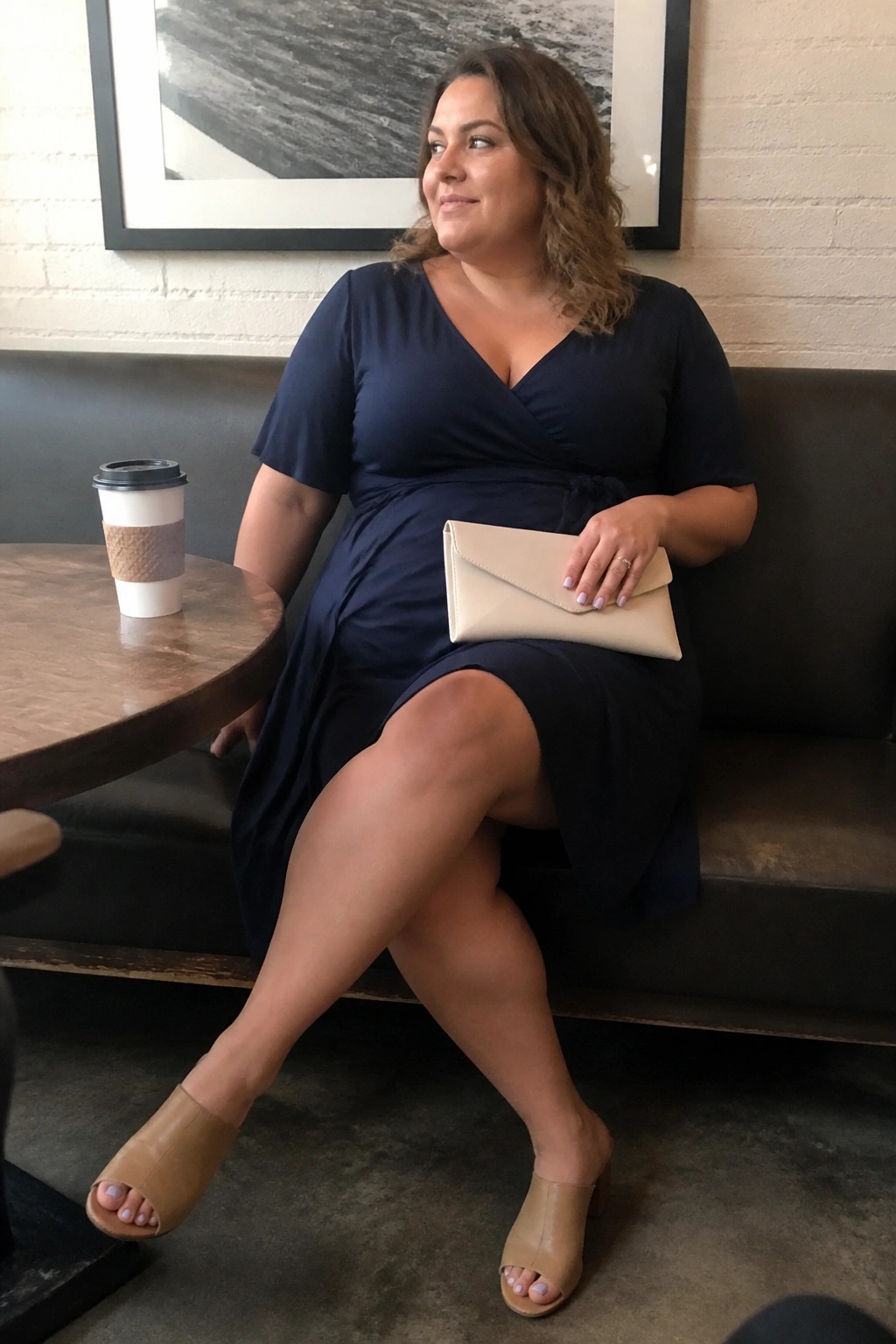 Woman in navy wrap dress holding beige clutch beside coffee on table