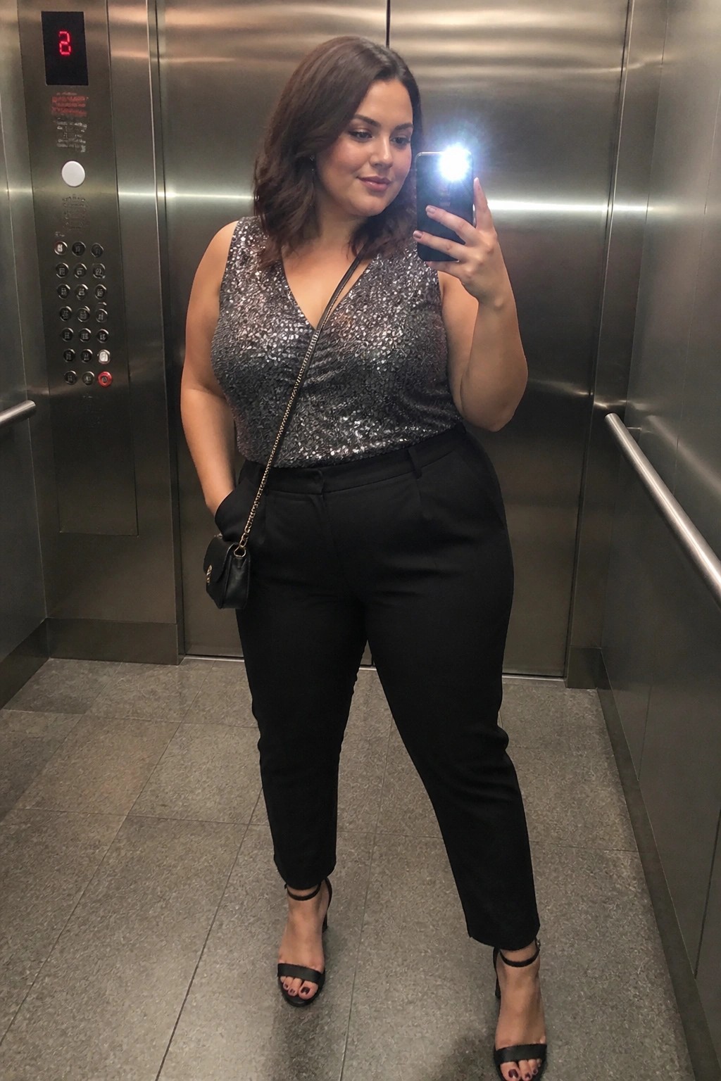 Woman in sparkly sequin top taking selfie inside stainless steel elevator with black handbag