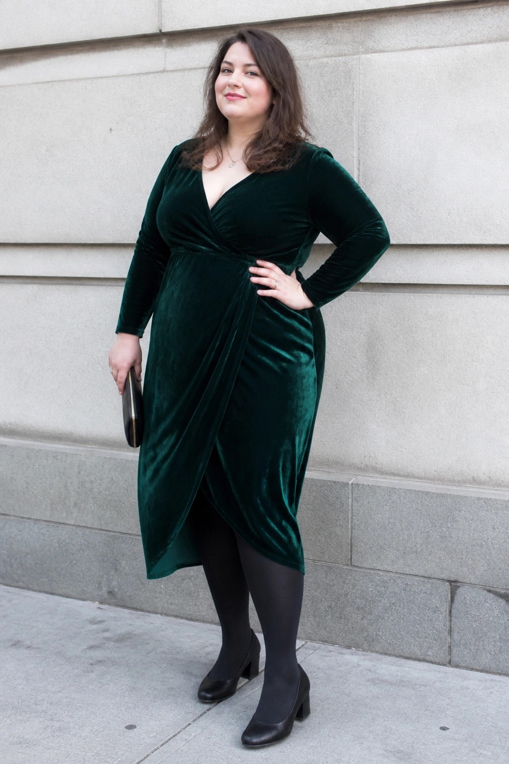 Woman in dark green velvet wrap dress with black tights and heels by stone wall