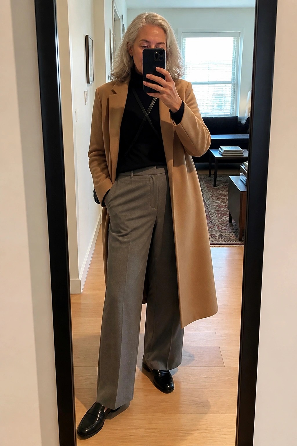 Woman over 60 wearing a long camel coat over a black turtleneck with gray wide-leg trousers and black loafers while taking a mirror selfie.