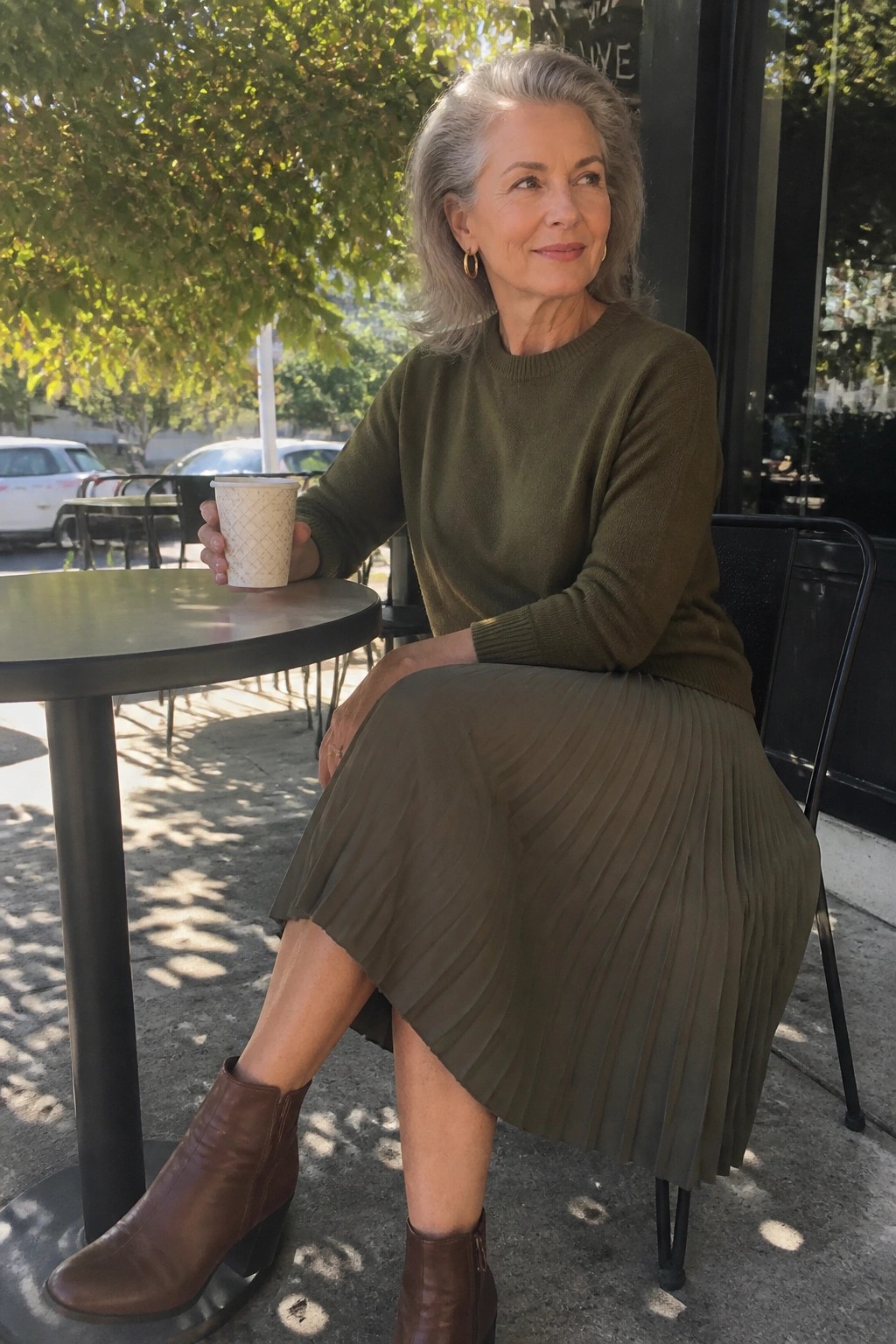 Gray-haired woman in olive sweater and pleated skirt holds coffee at sunny outdoor table.