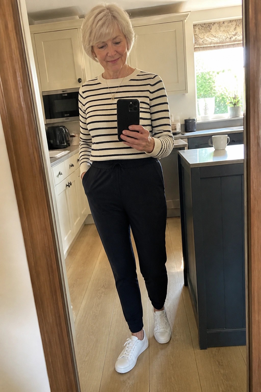 Woman in striped sweater and black pants taking mirror selfie in kitchen