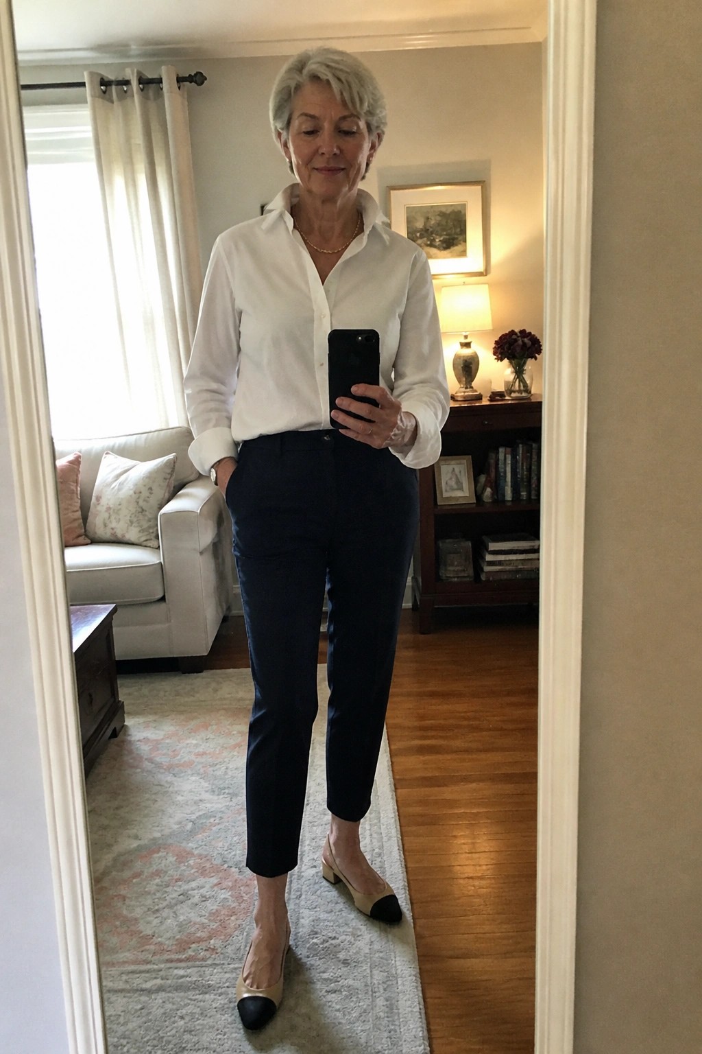 Gray-haired woman in white shirt and navy pants taking mirror selfie with phone