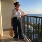 Woman wearing a white linen button-down shirt tied in a front knot at the waist paired with navy wide-leg linen pants, brown leather slide sandals, and holding a woven straw tote bag.