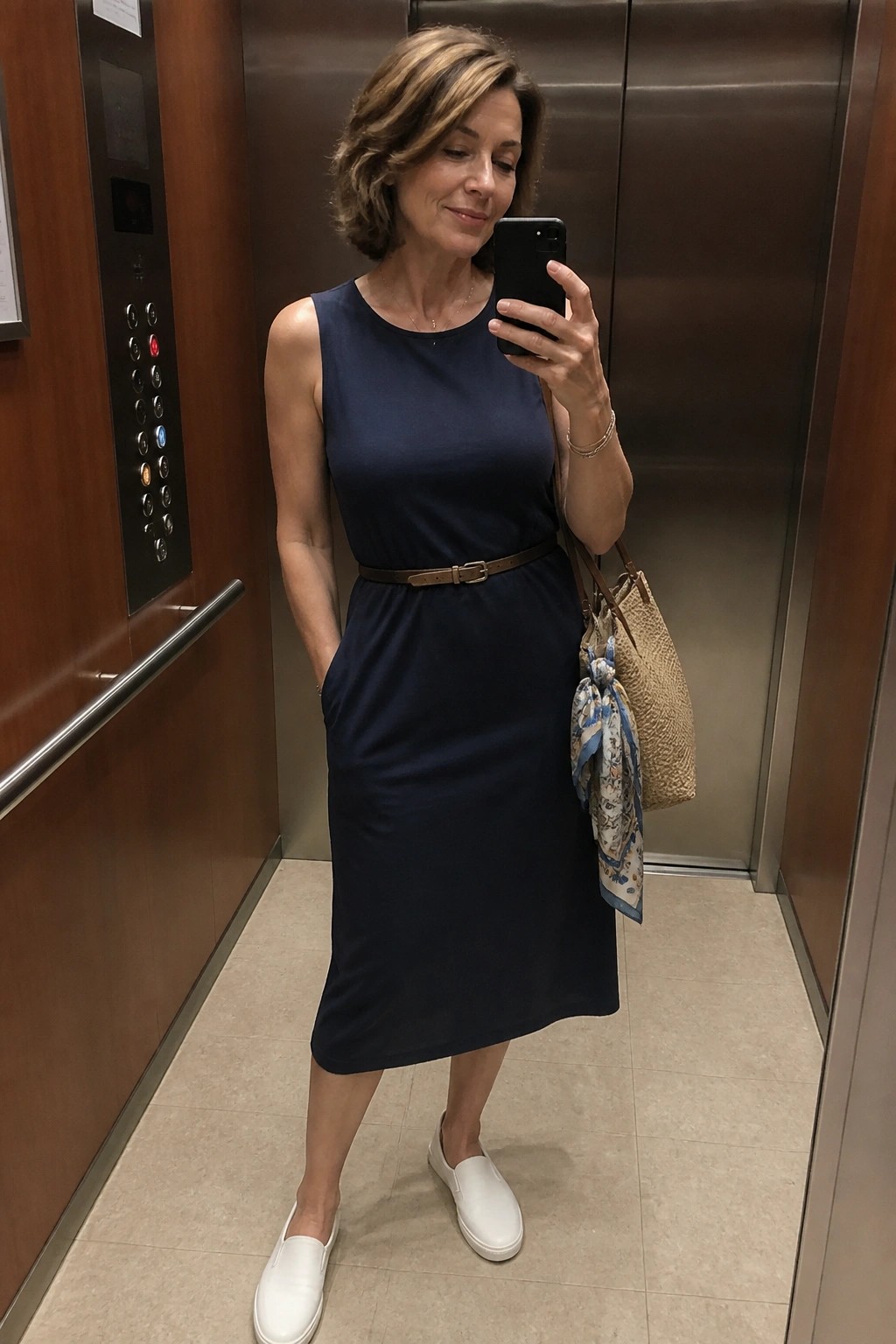 Woman in navy dress taking selfie in elevator with straw handbag