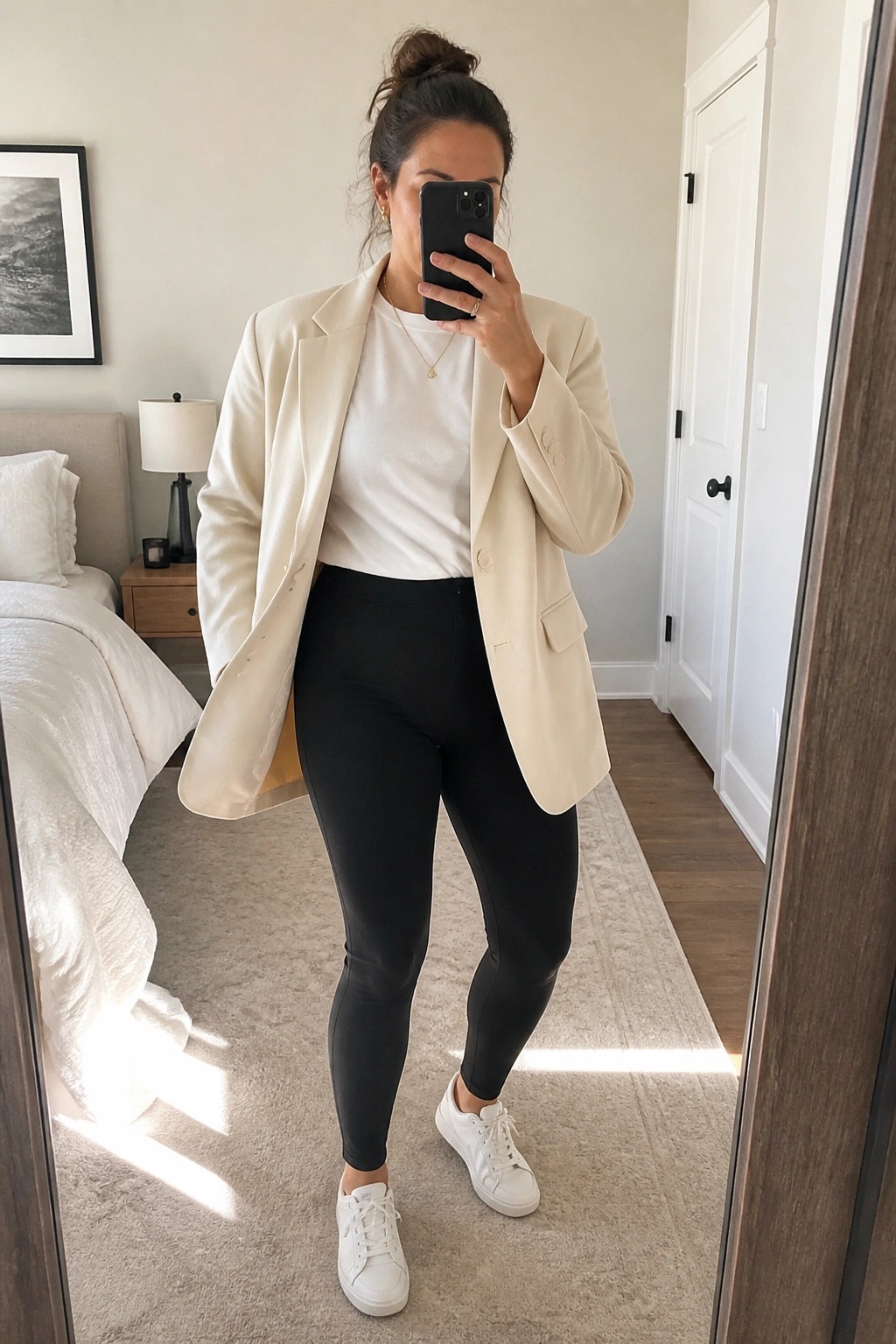 Woman wearing an open cream blazer over a white t-shirt with black leggings and white sneakers, standing in a bedroom while holding a phone.