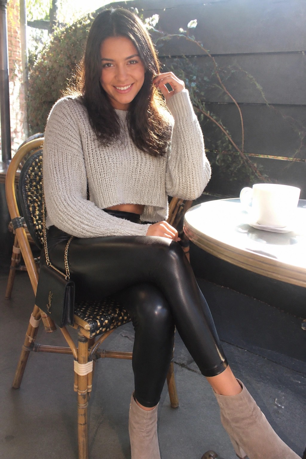 Woman in gray cropped sweater and black leggings at sunny outdoor cafe table