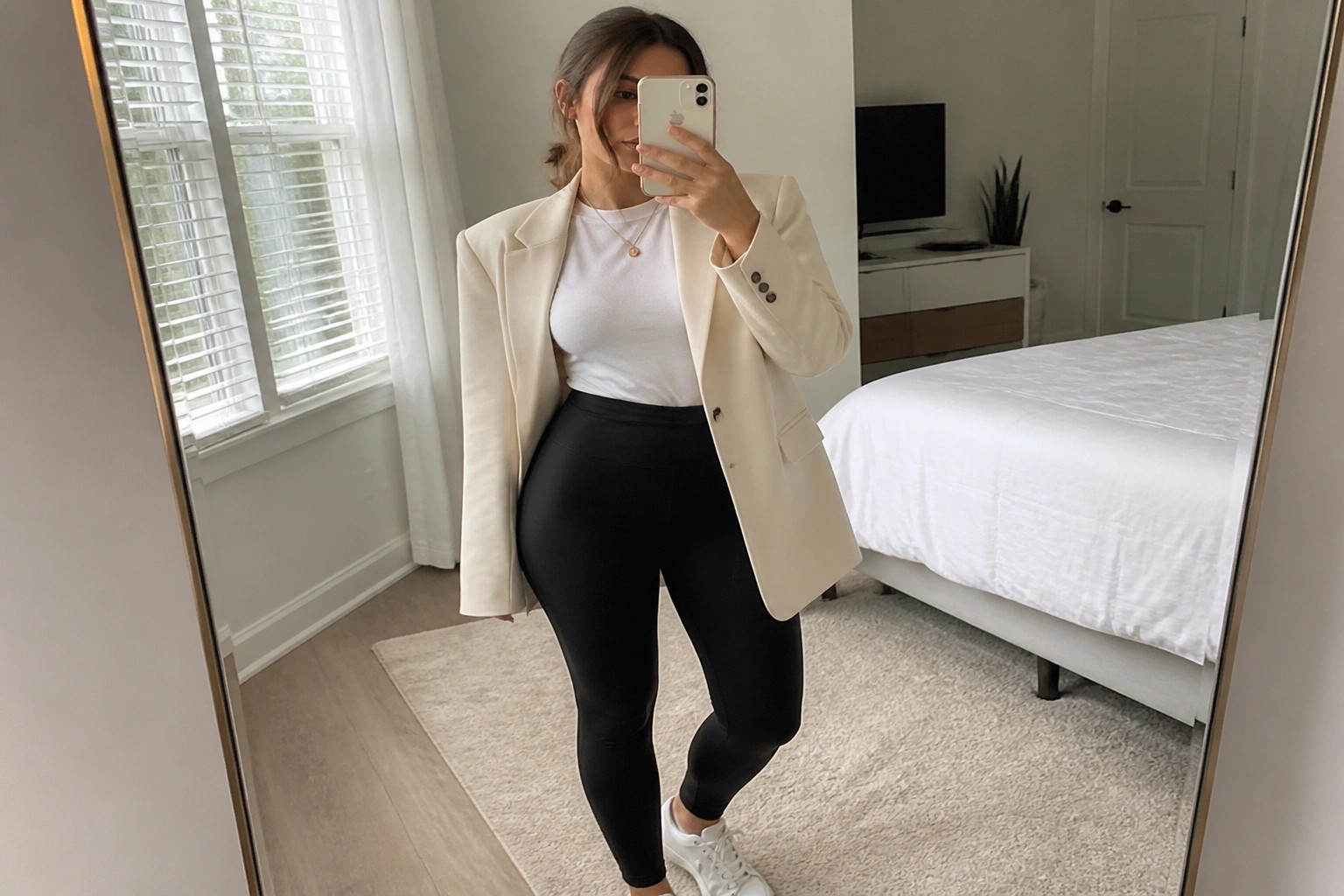 Woman wearing an open cream blazer over a white t-shirt with black leggings and white sneakers, standing in a bedroom while holding a phone.